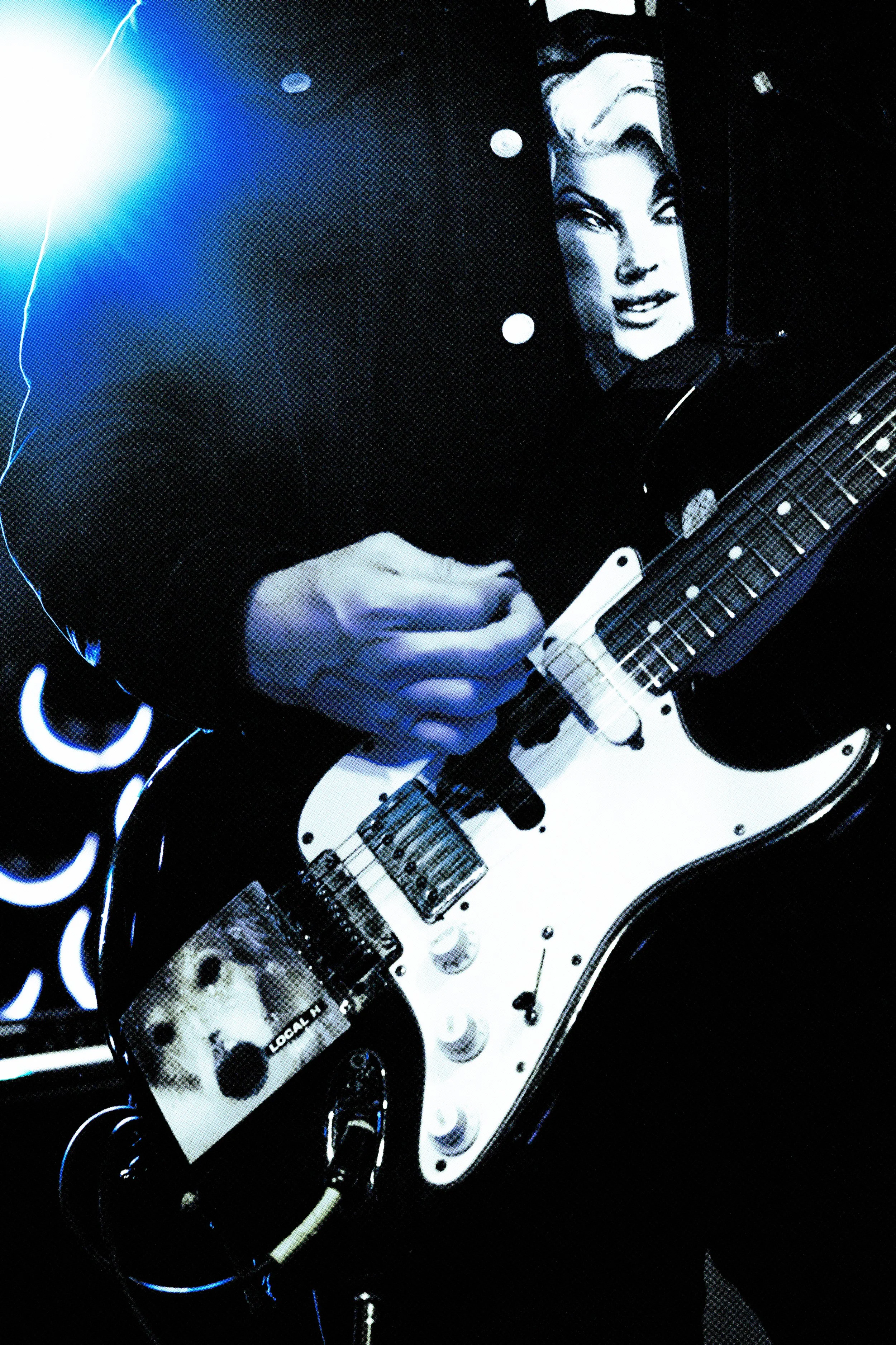 Person playing an electric guitar wearing a black jacket with a picture of a woman on it, in a dark setting with blue lighting.