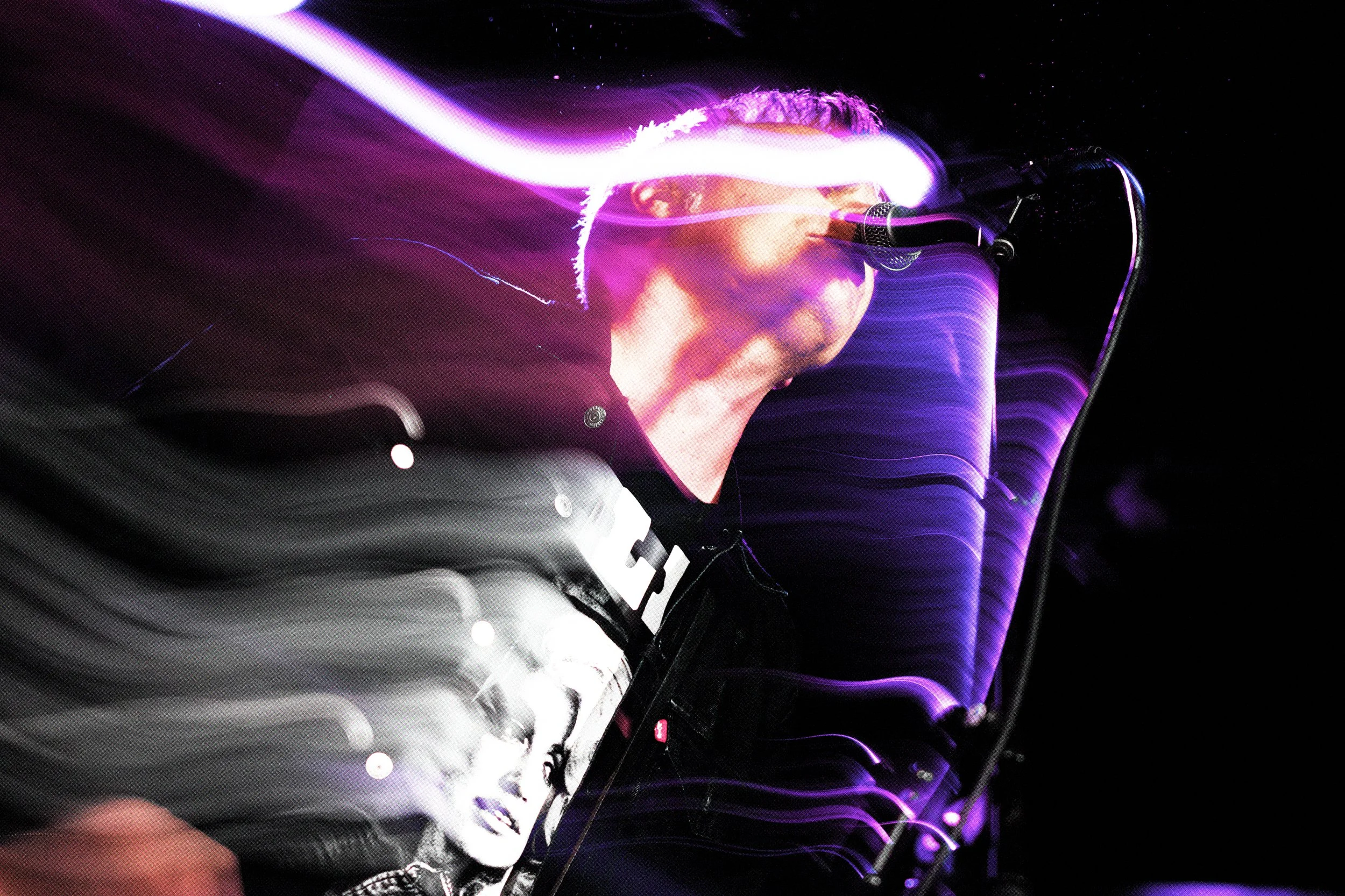 A person wearing glasses with long hair, illuminated by purple and white lights, performing with a microphone in front of a screen displaying a woman's face.