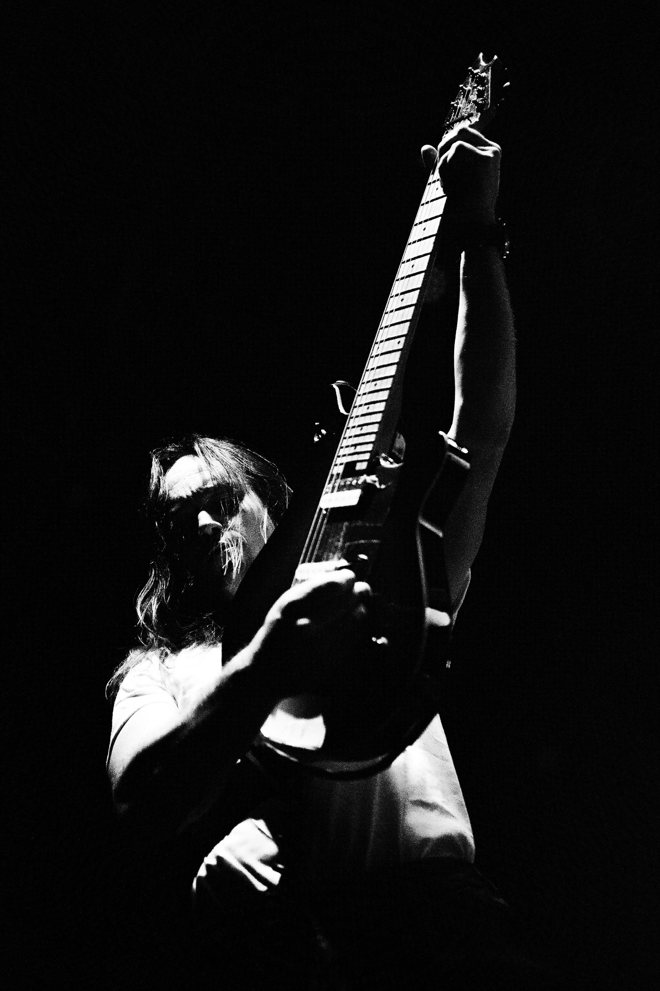 A person holding an electric guitar, viewed from a low-angle shot in a dark setting, with dramatic lighting.