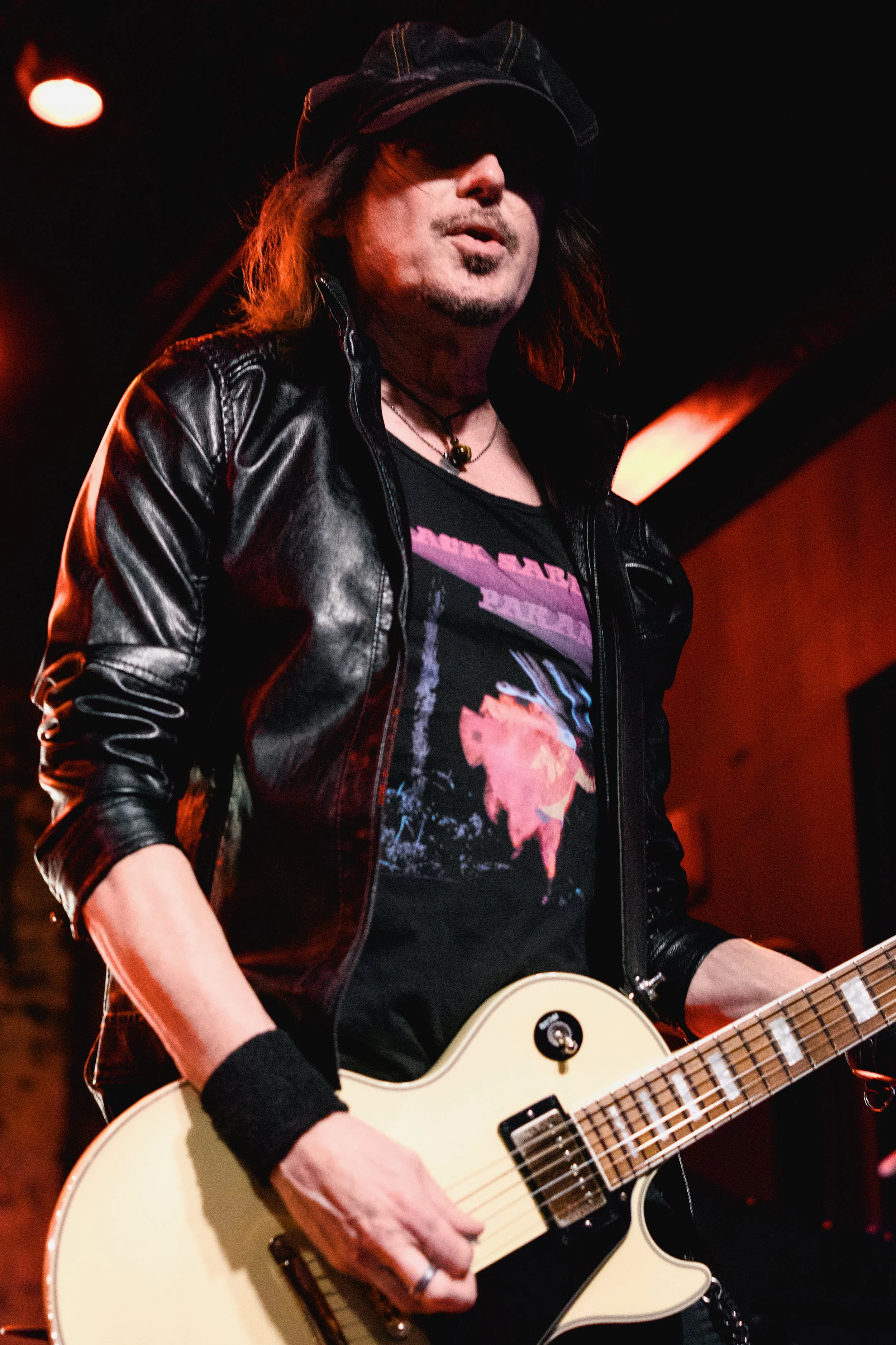A male musician playing an electric guitar on stage, wearing a black leather jacket, black graphic T-shirt, hat, and wristband, with red stage lighting.