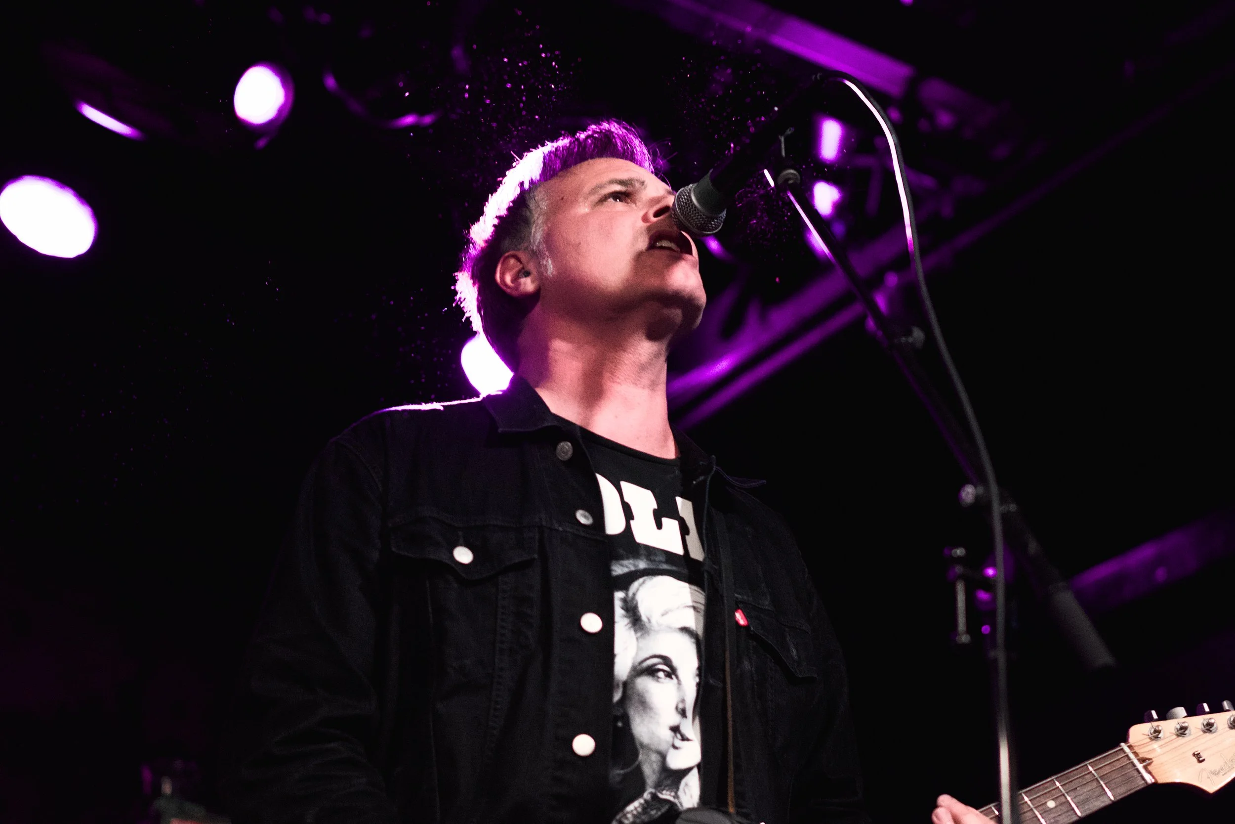 A musician performing on stage, singing into a microphone with purple stage lights in the background, wearing a black jacket and a graphic T-shirt.