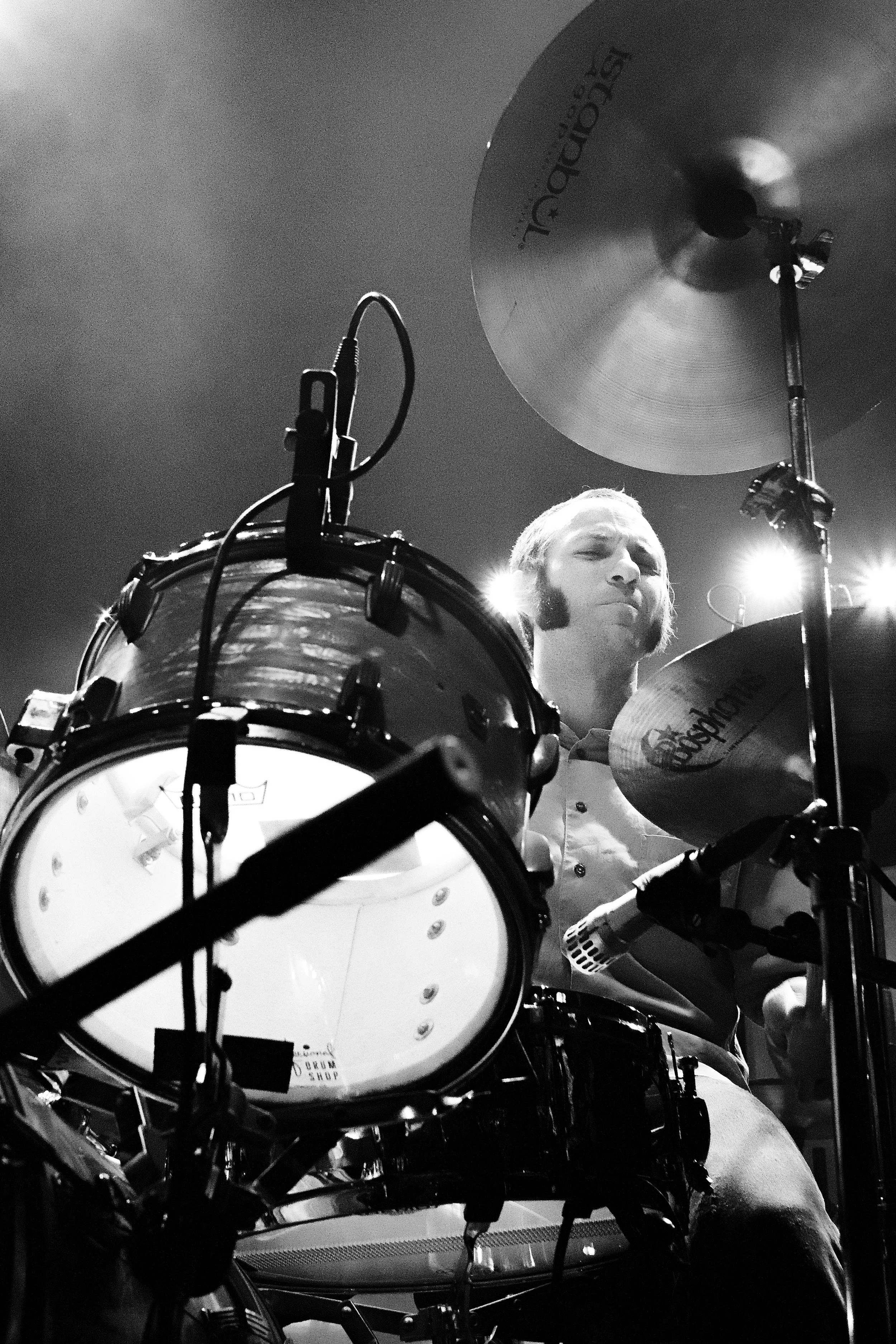 Black and white photo of a drummer playing his drum kit on stage, with bright stage lights behind him.