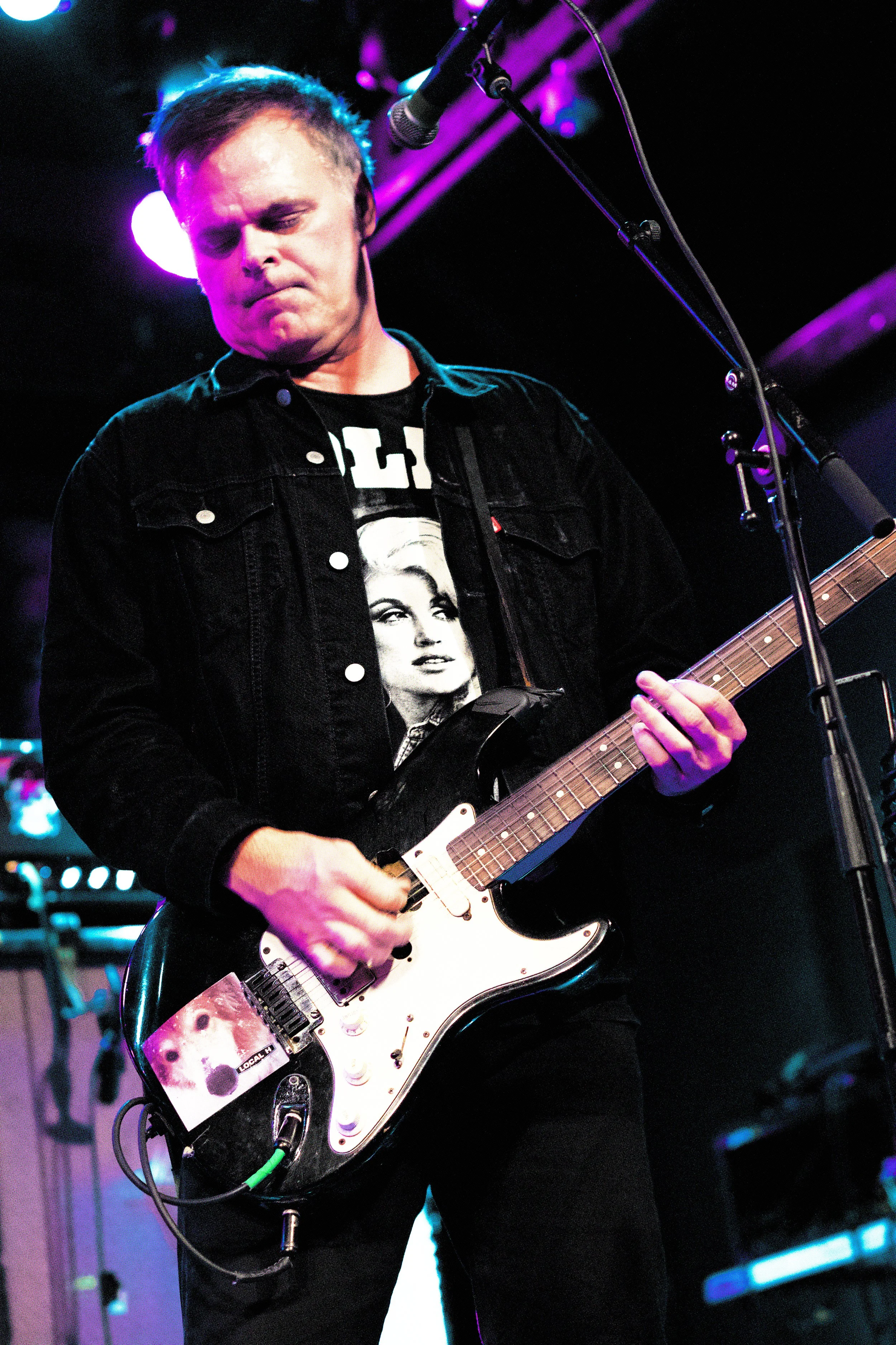 A man playing an electric guitar on stage, wearing a black denim jacket and a T-shirt with a woman's face on it, with purple and blue stage lights in the background.