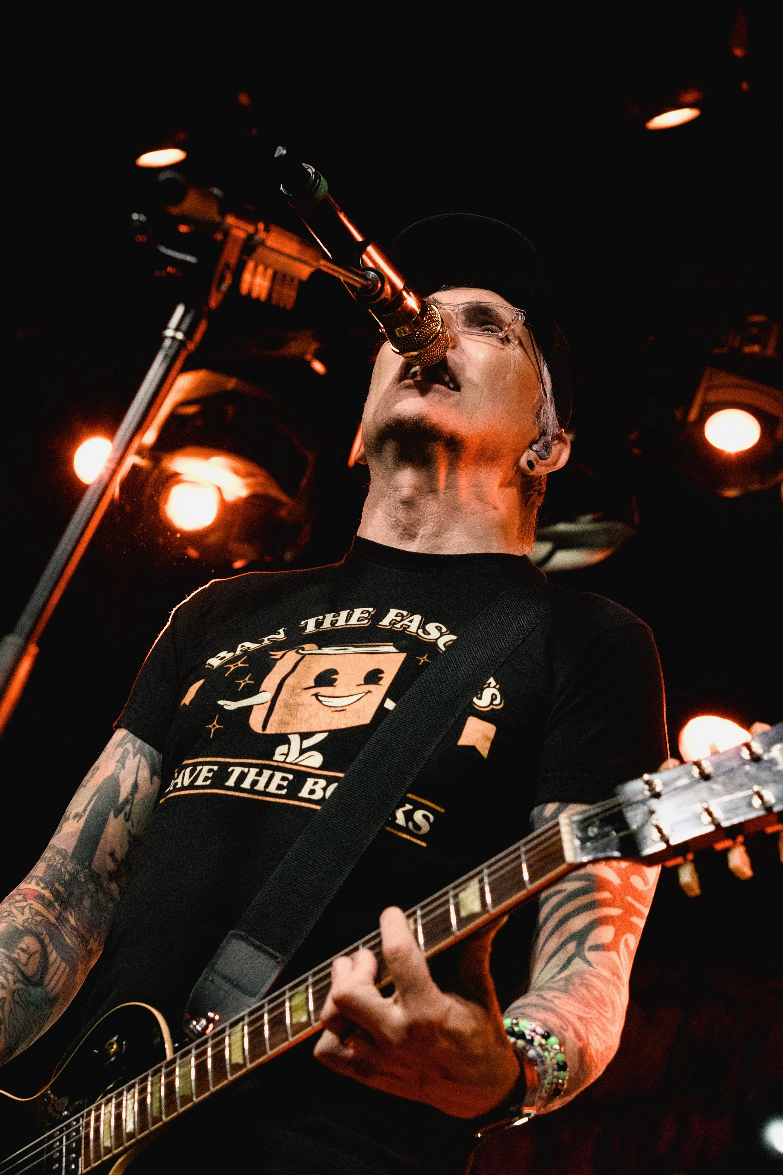 A male musician singing into a microphone while playing an electric guitar on stage, with tattoos on his arms and wearing glasses, a black t-shirt, and a cap, under stage lighting.