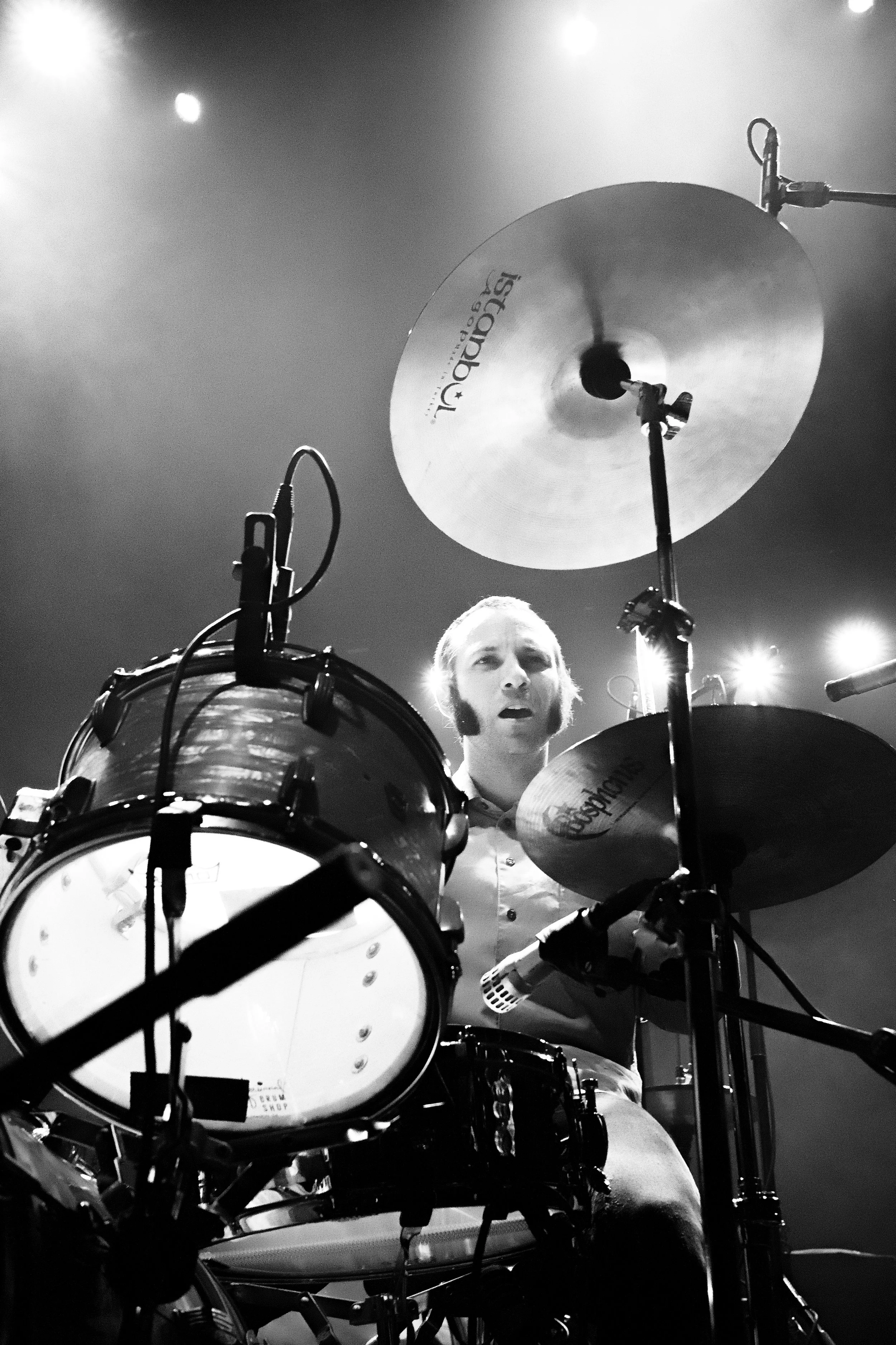 A black and white photo of a drummer performing on stage, surrounded by drums and cymbals, with stage lights in the background.