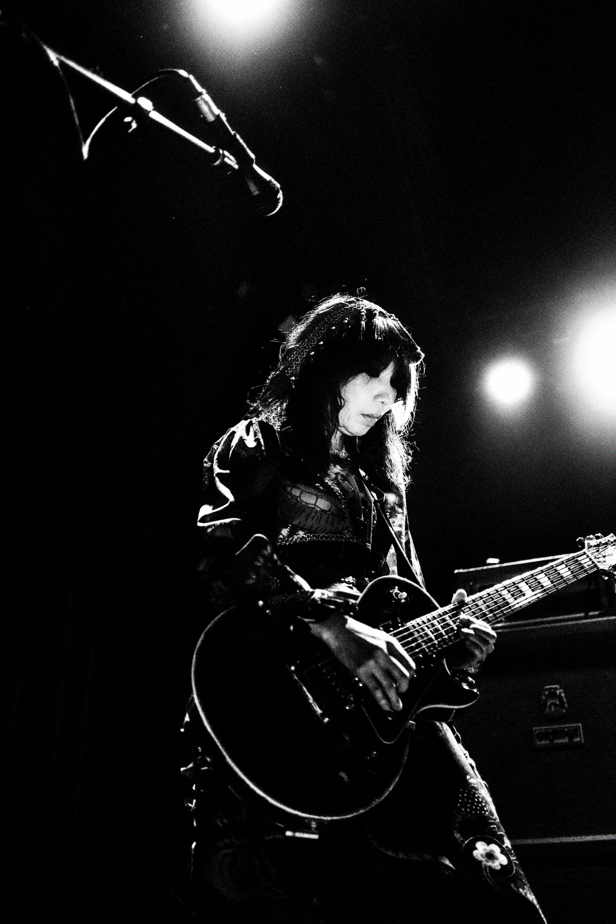 A female musician playing an electric guitar on stage with bright lights behind her in black and white.