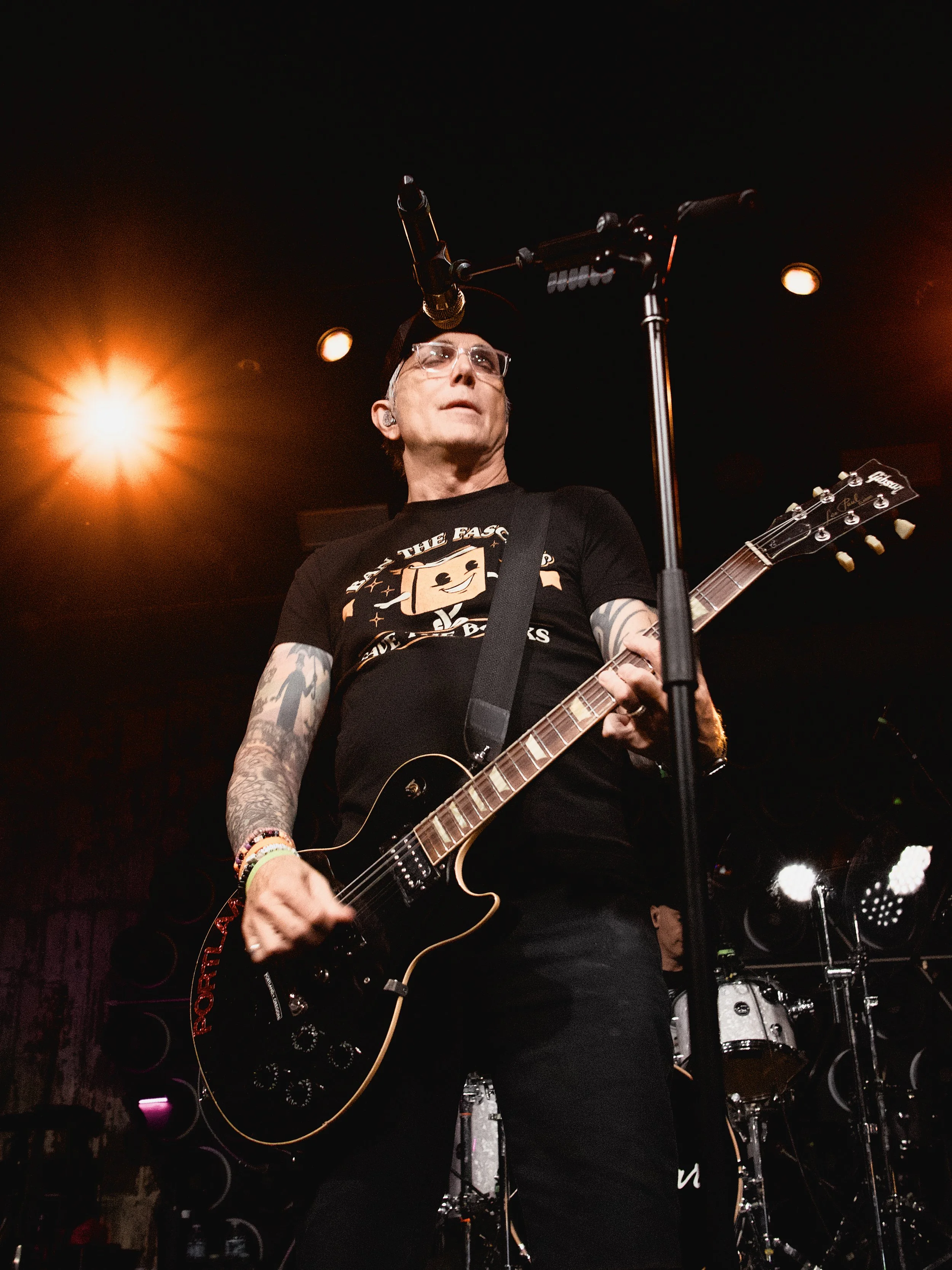 A man wearing sunglasses and a black T-shirt playing an electric guitar on stage, with microphones and stage lights overhead.