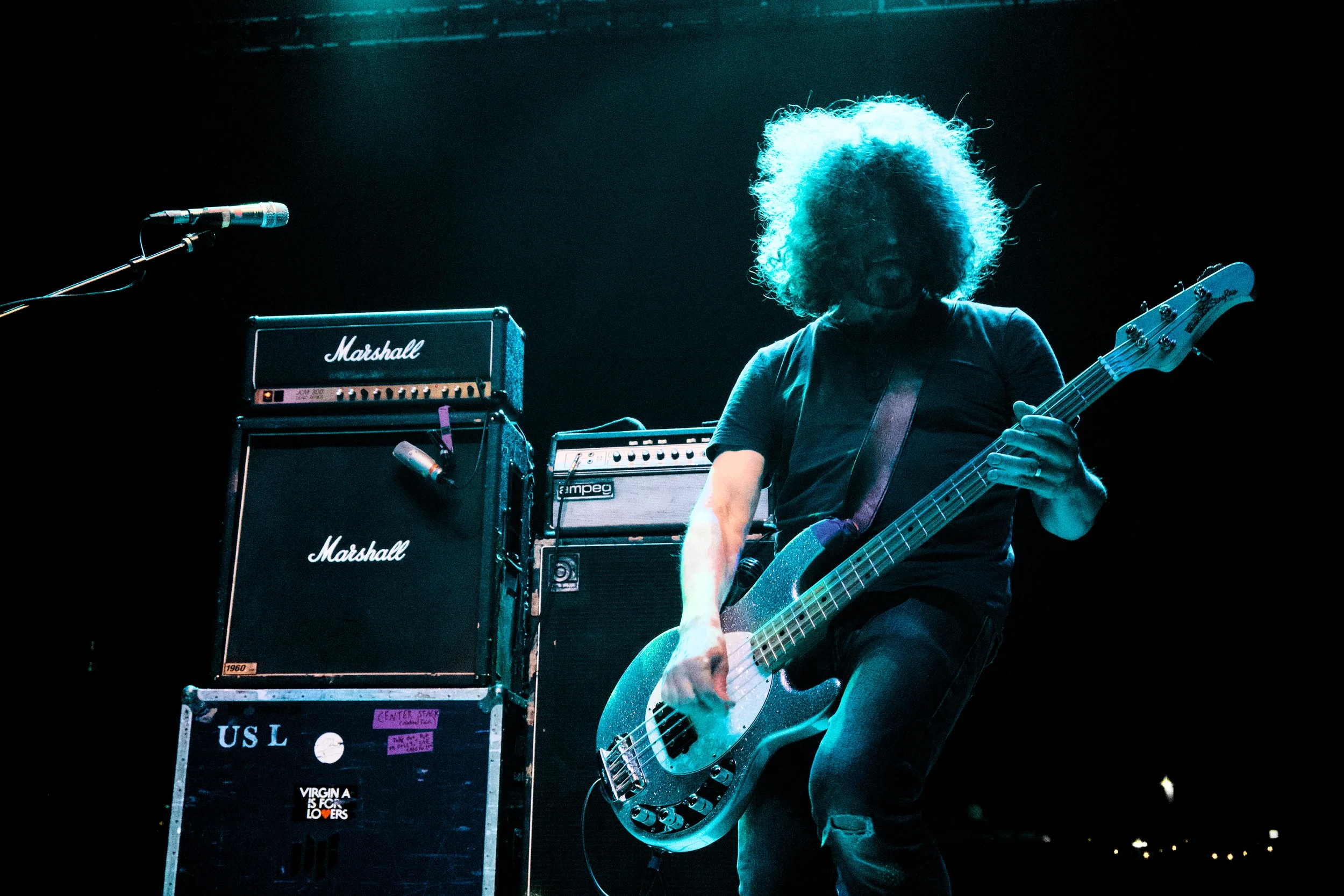 A musician with curly hair playing an electric bass guitar on stage, with Marshall amplifiers in the background, lit by blue stage lighting.