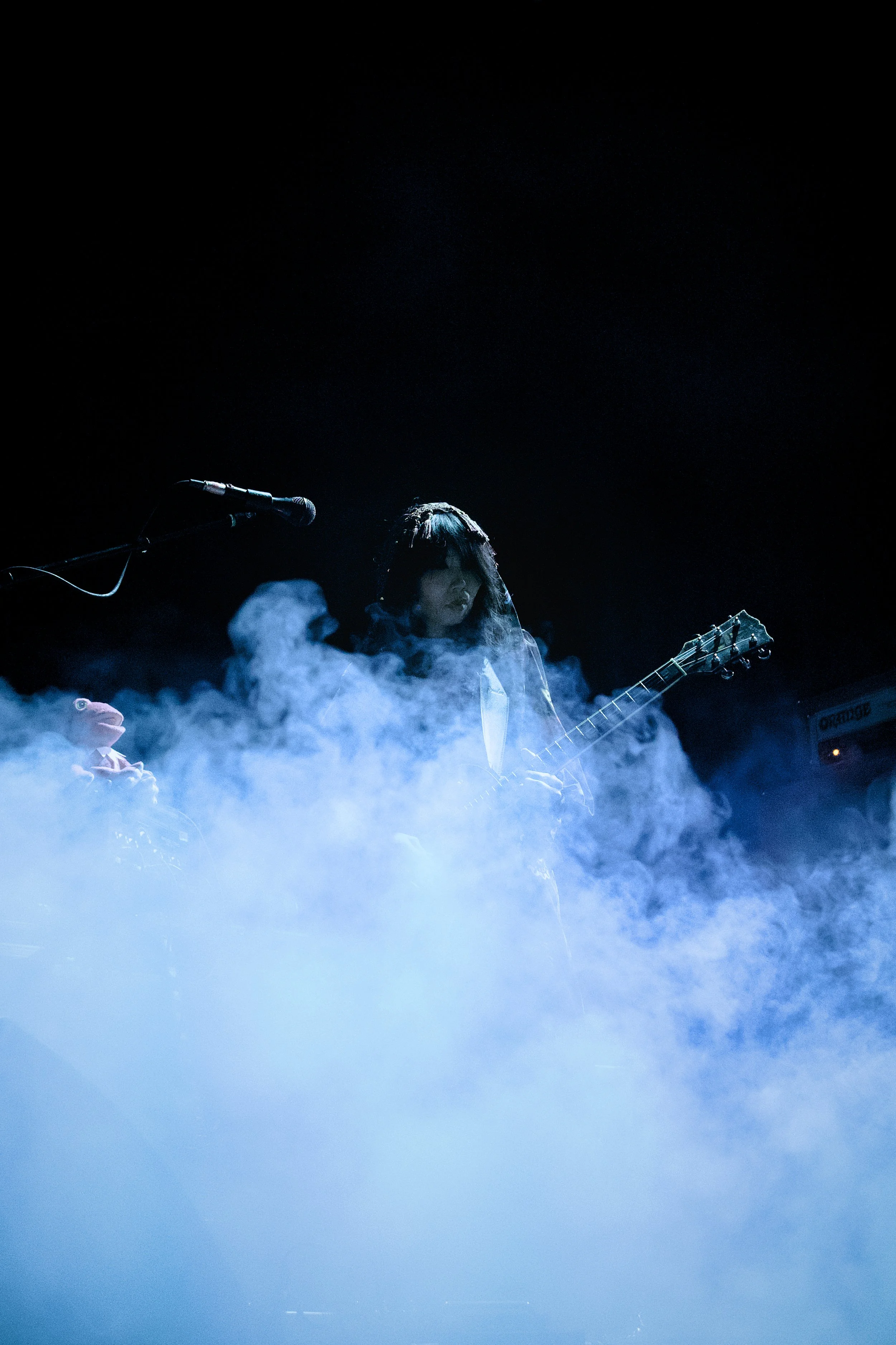 A female musician playing an electric guitar during a performance with stage fog and a dark background.