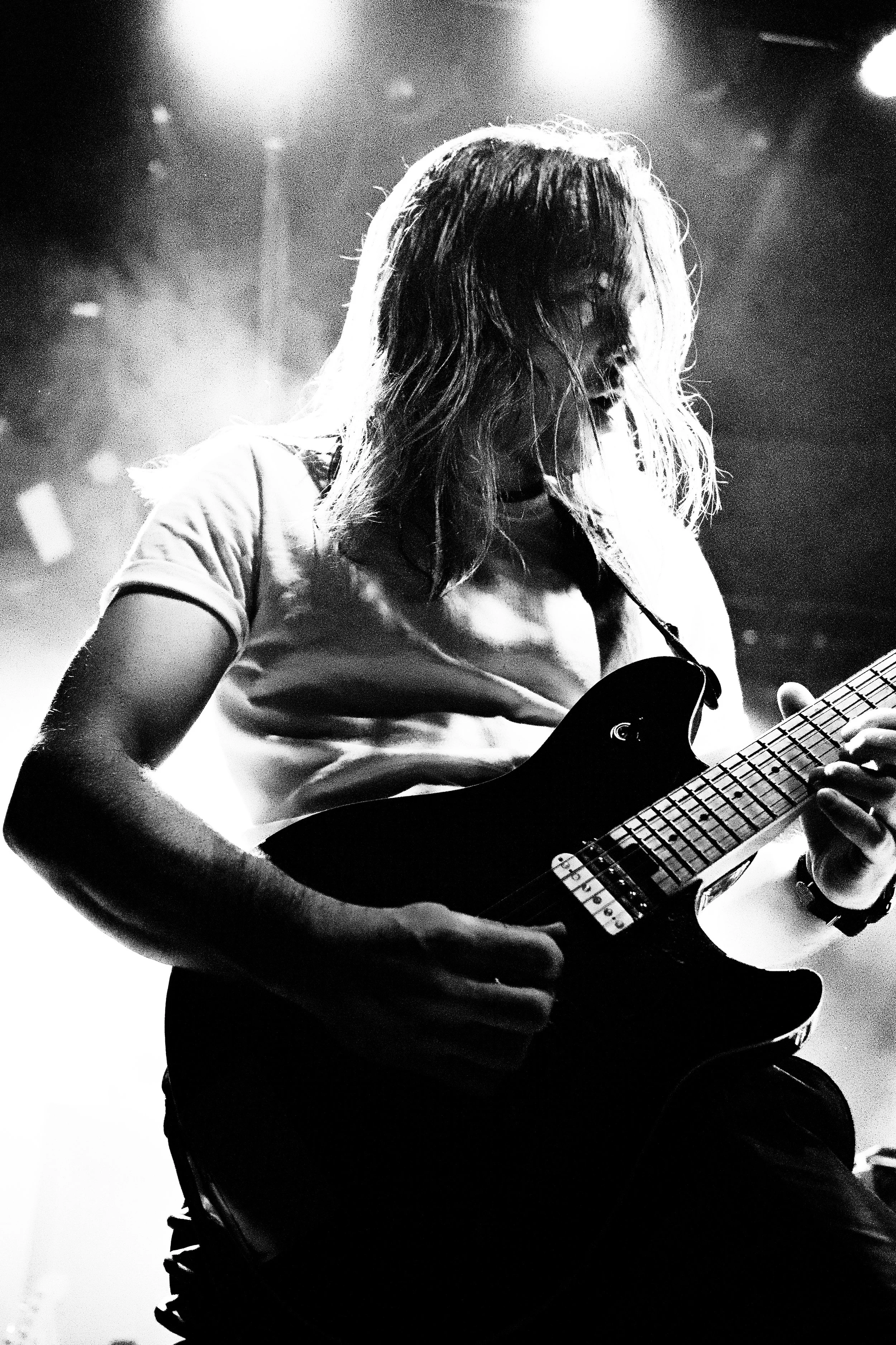 Black and white photo of a woman playing an electric guitar on stage, with her hair covering part of her face and stage lights illuminating the scene.