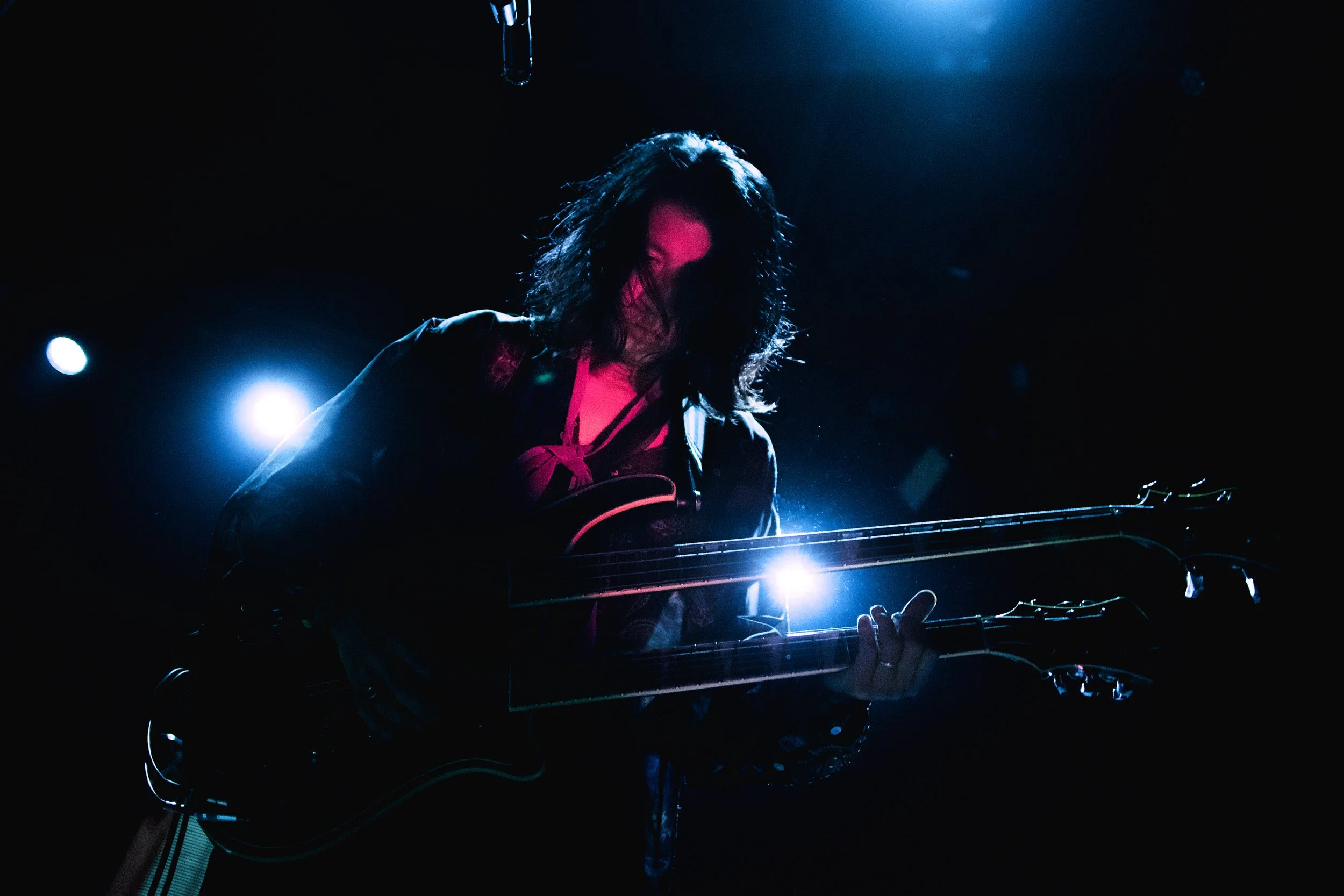 A musician with long hair playing an electric guitar on a dark stage illuminated by blue and pink lights.