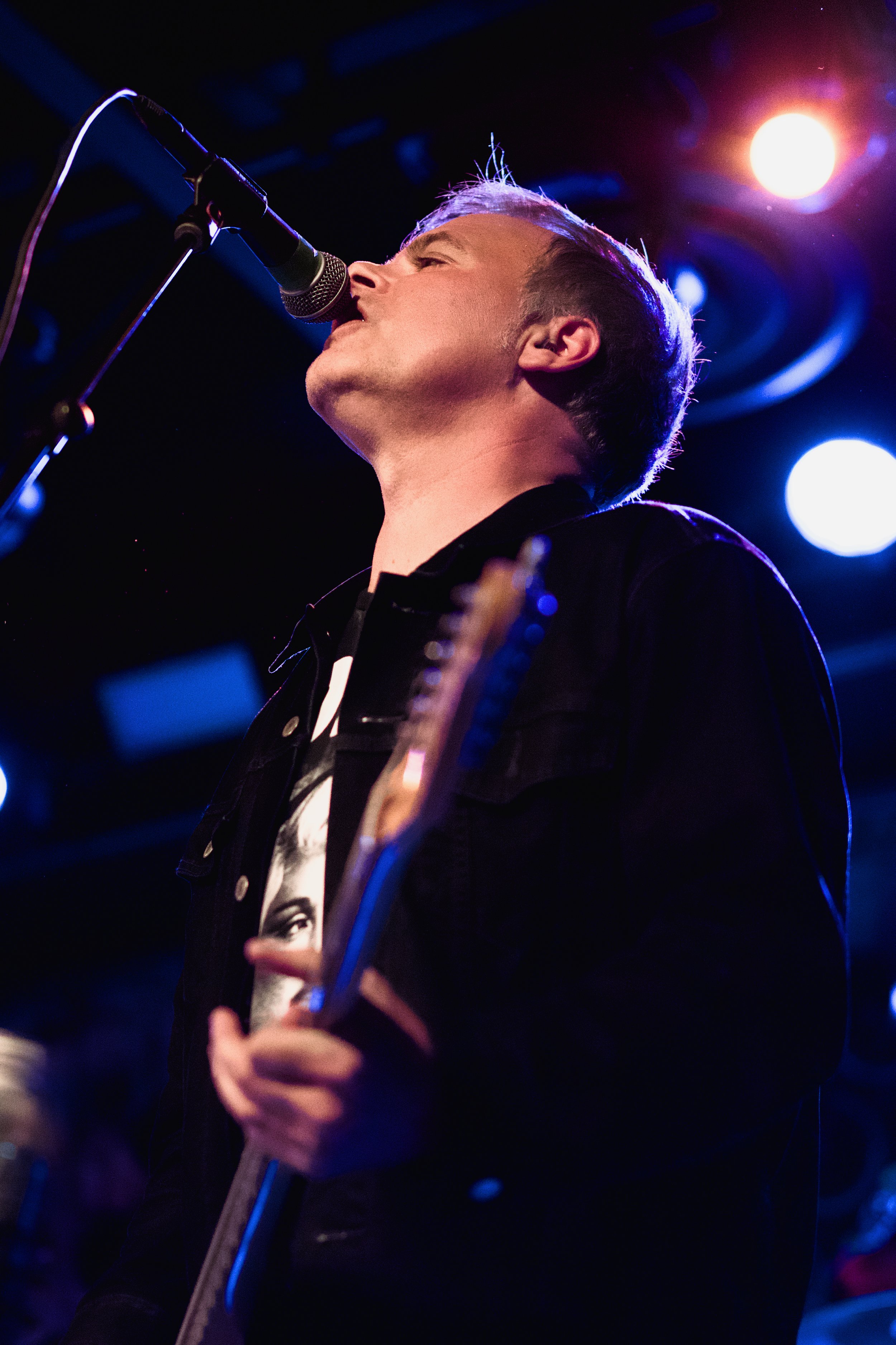 A male musician singing into a microphone on stage with colorful lights in the background.