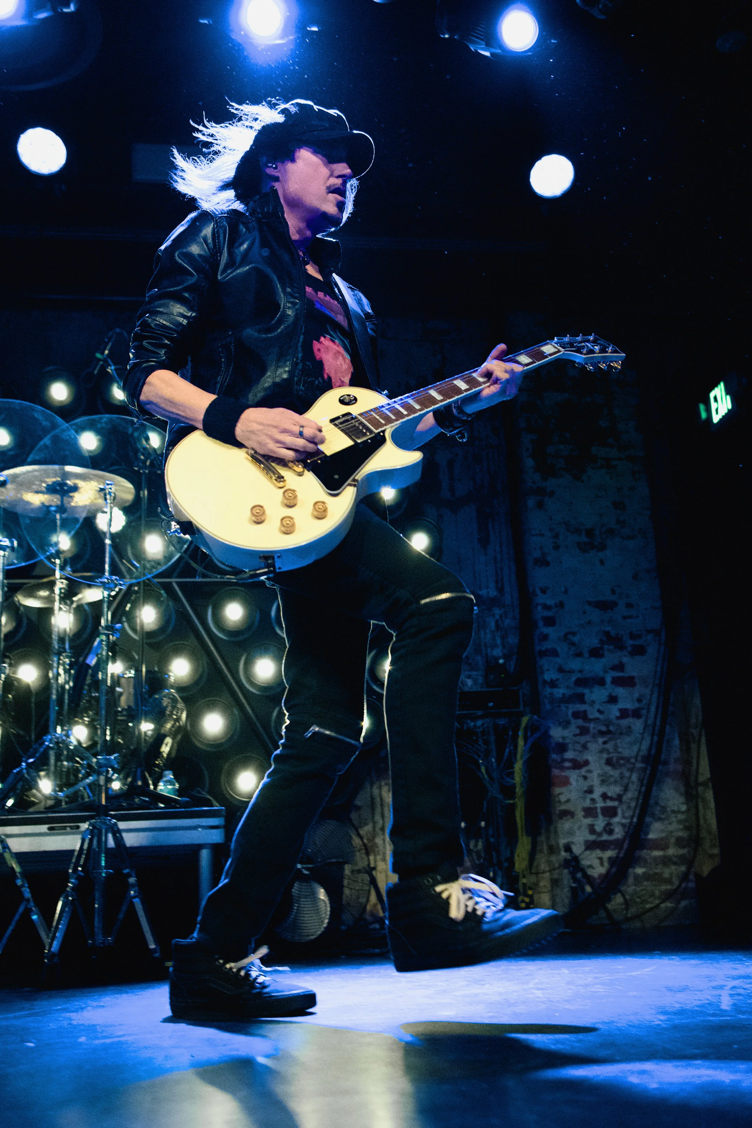 A woman playing an electric guitar on stage at a concert, with a drum set and stage lights in the background.