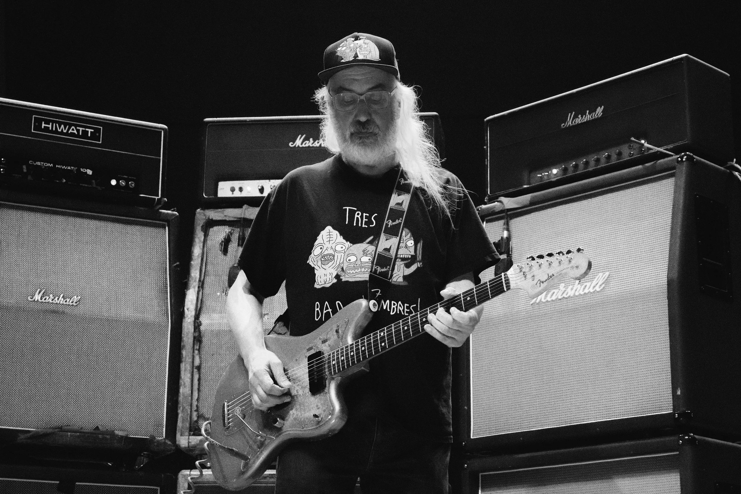 A man with long hair, beard, glasses, wearing a cap and a black T-shirt with cartoon skulls, playing an electric guitar in a room with Marshall and Hiwatt amplifiers.