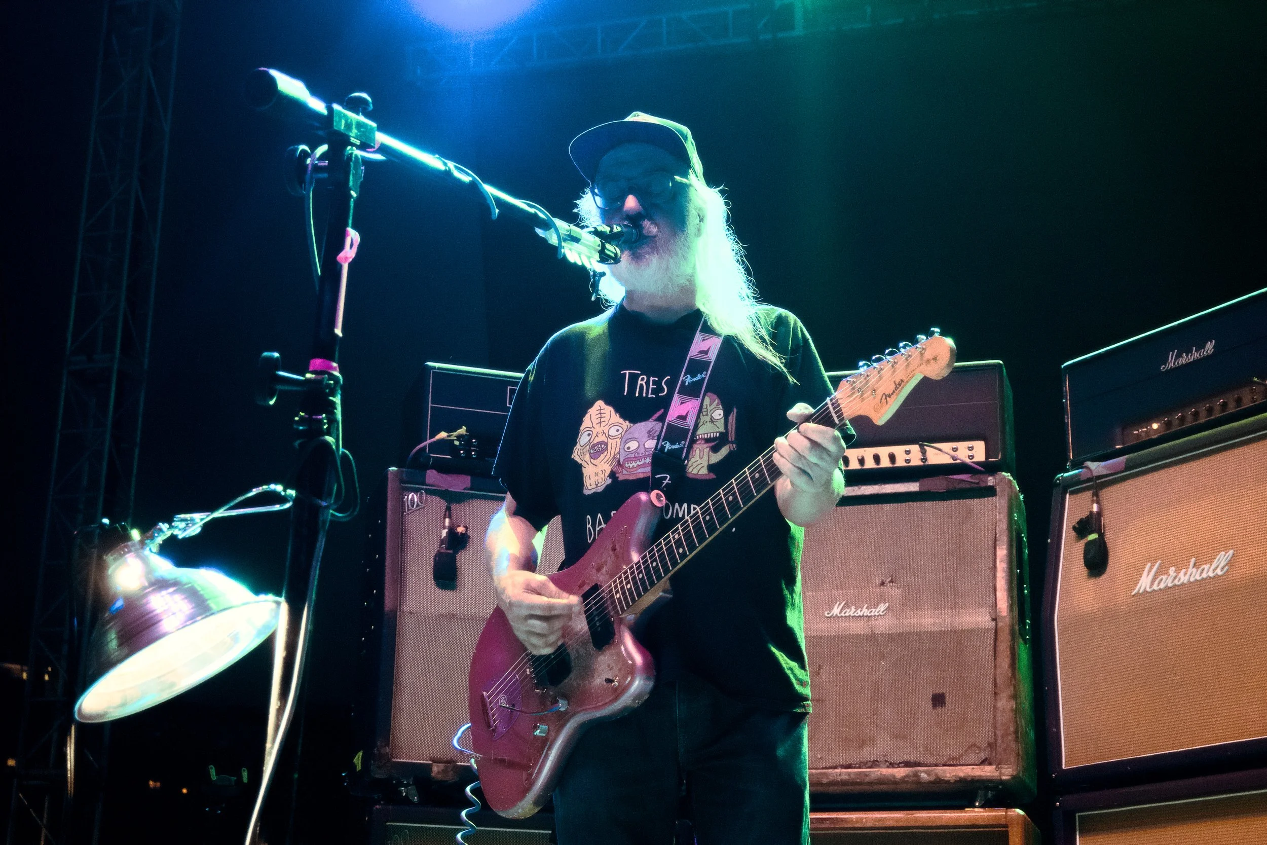 A man with a long white beard, glasses, and a cap playing an electric guitar on stage with a microphone in front of him. Behind him are multiple Marshall amplifiers.
