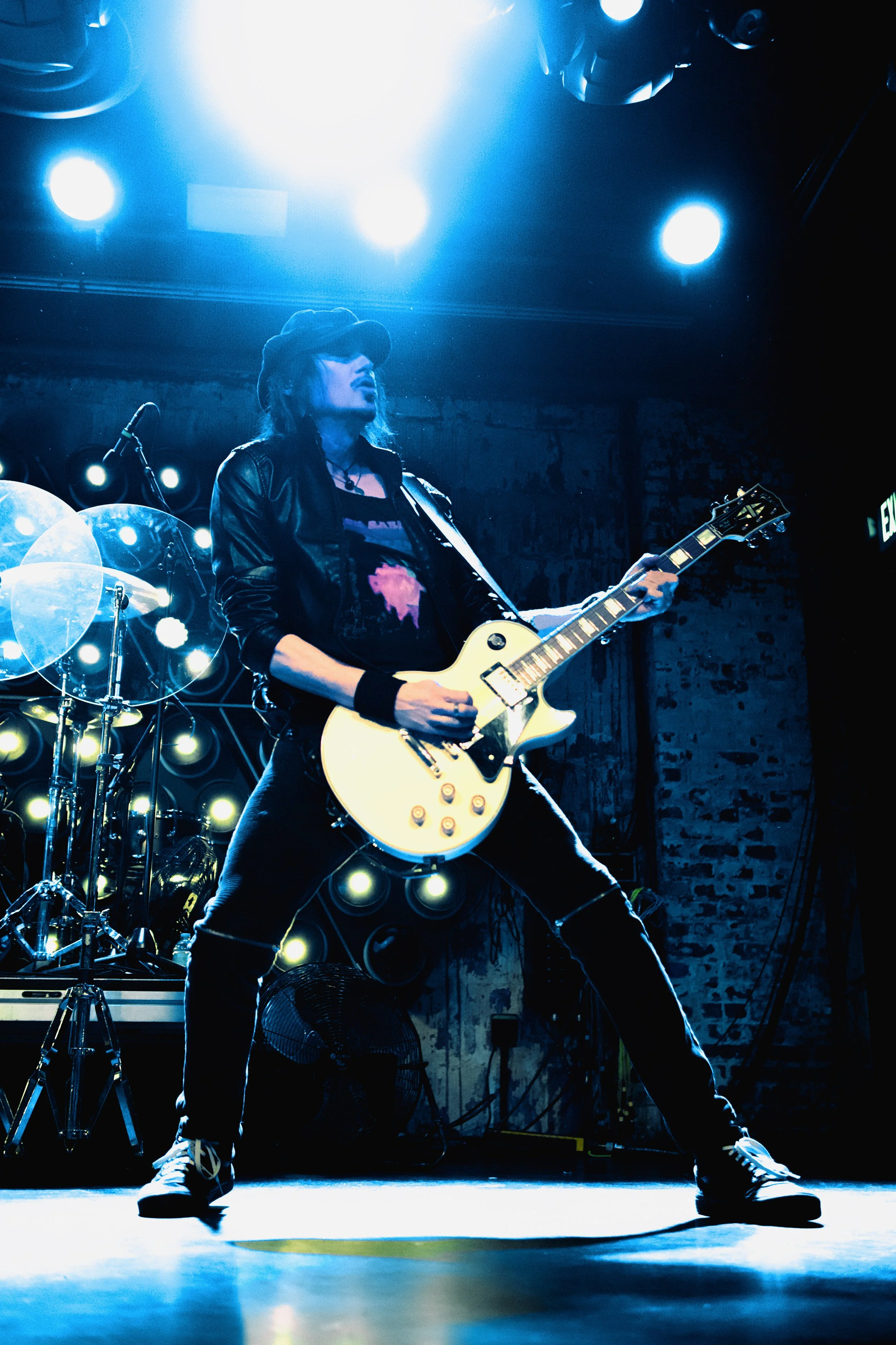 A female guitarist performing on stage during a concert, illuminated by blue stage lights, wearing a black leather jacket, a cap, and jeans.