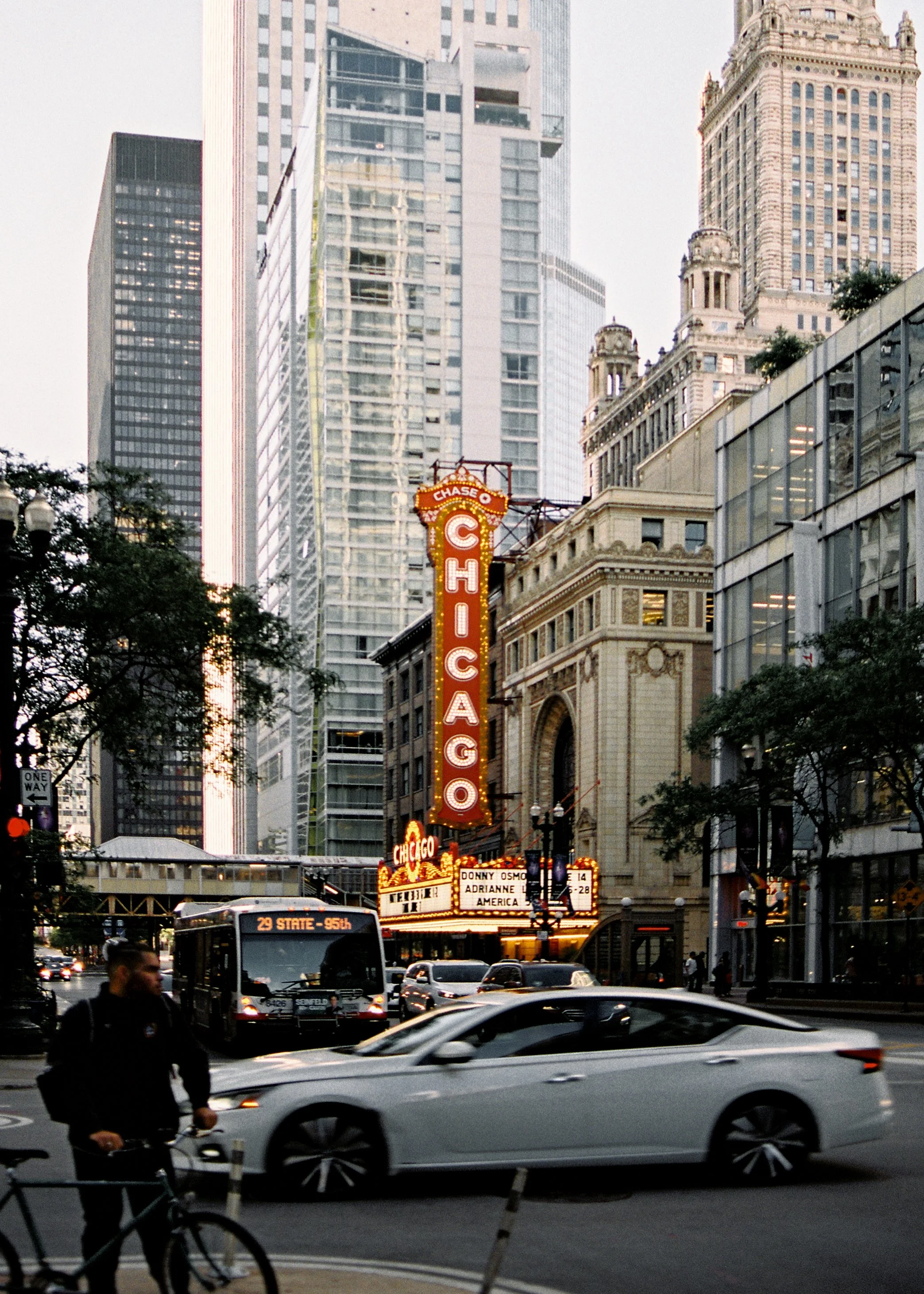 Street scene in Chicago with the iconic marquee sign of the Chicago Theatre, surrounded by tall modern and historic buildings, with cars and a cyclist in the foreground.