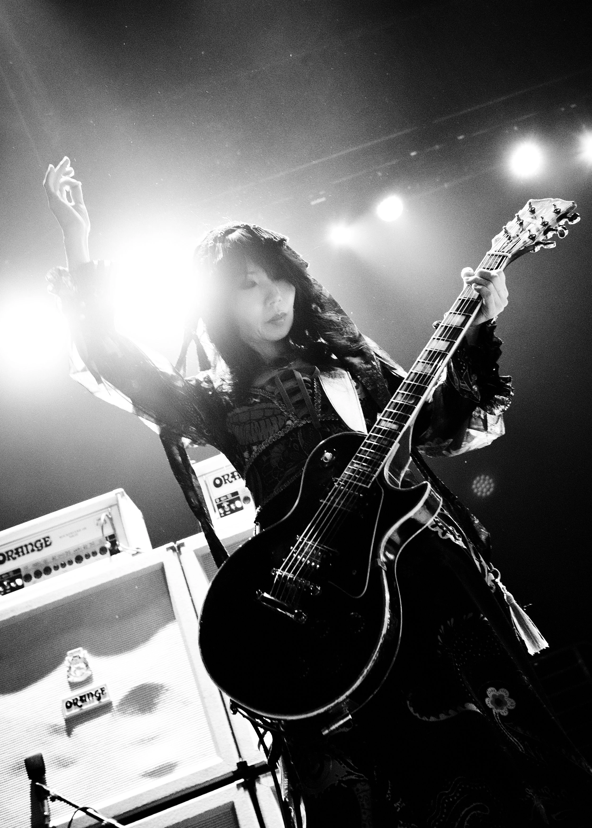 A female musician playing an electric guitar on stage, surrounded by amplifiers and bright stage lights.