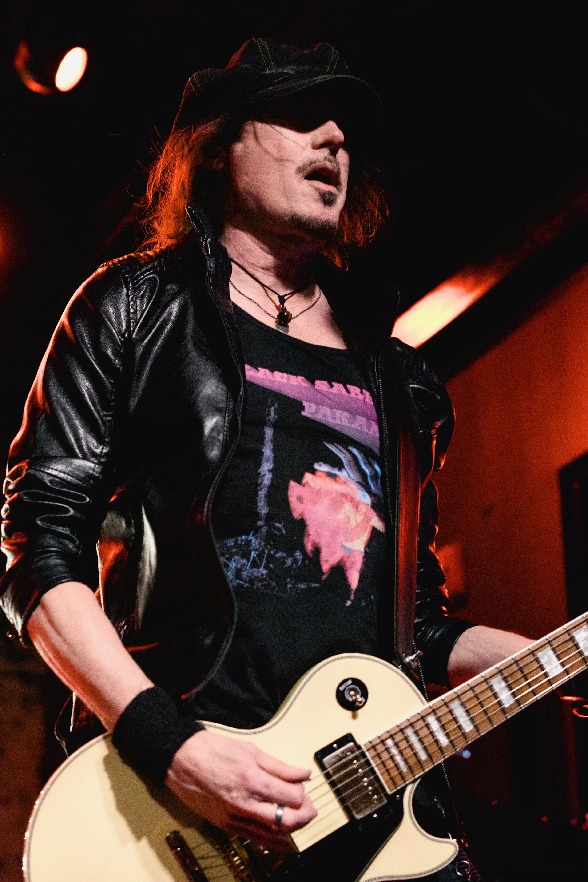 A male musician performing on stage, wearing a black leather jacket, a black graphic T-shirt, a black cap, and a necklace, playing a cream-colored electric guitar during a live concert under red stage lighting.