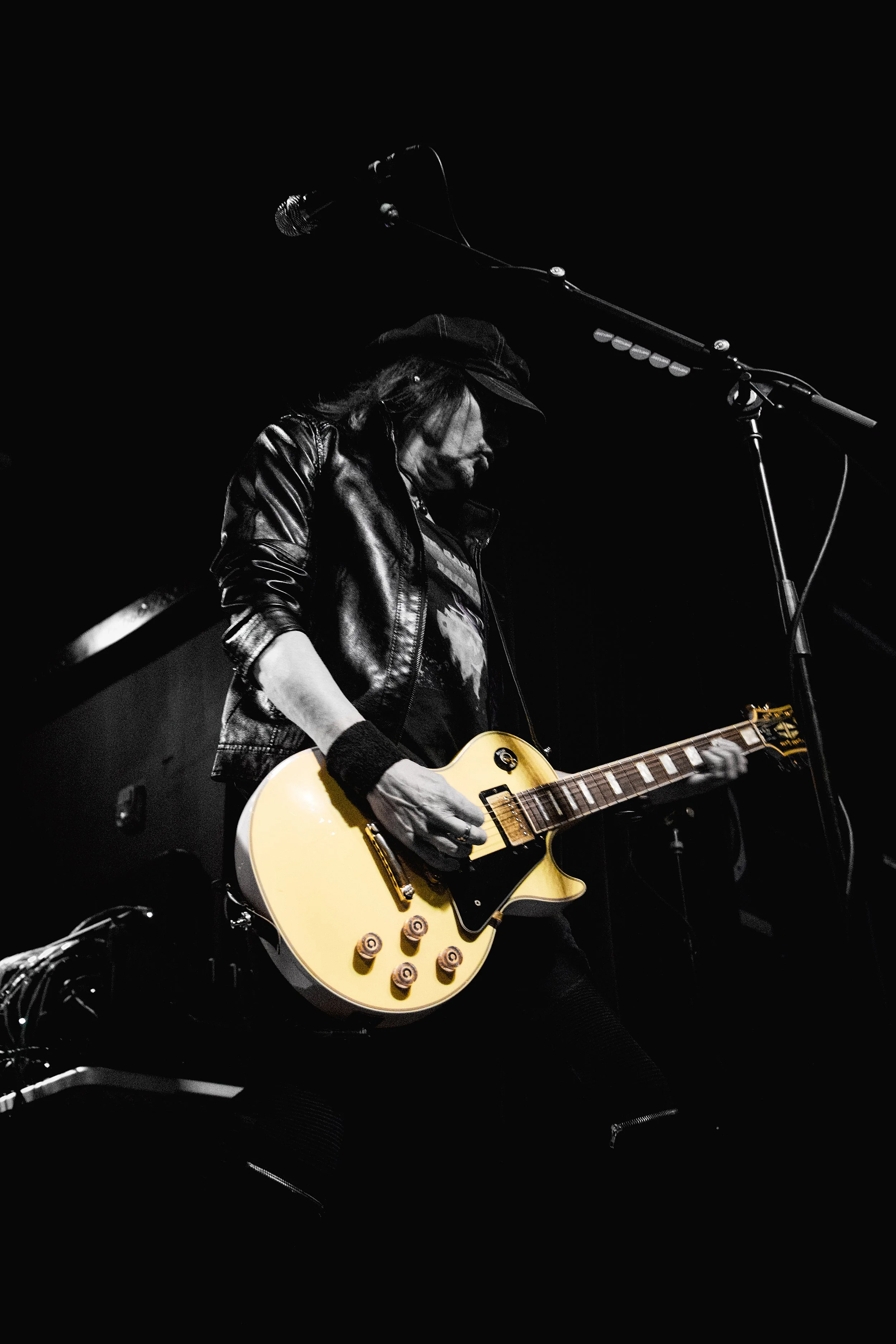 A musician playing an electric guitar on stage, wearing a leather jacket and a hat, with a microphone overhead.