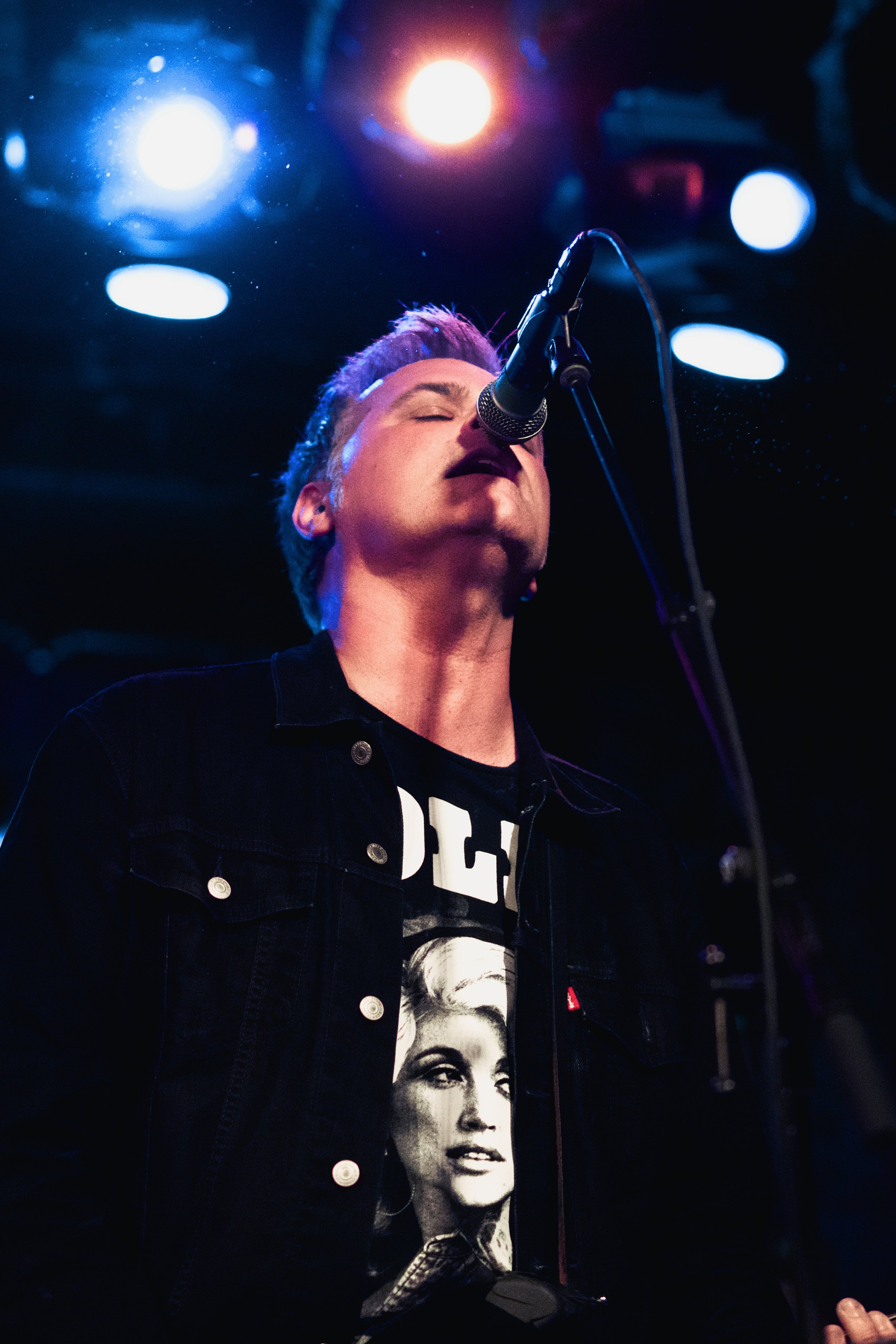Male singer performing on stage with closed eyes, standing in front of a microphone, illuminated by colorful stage lights.