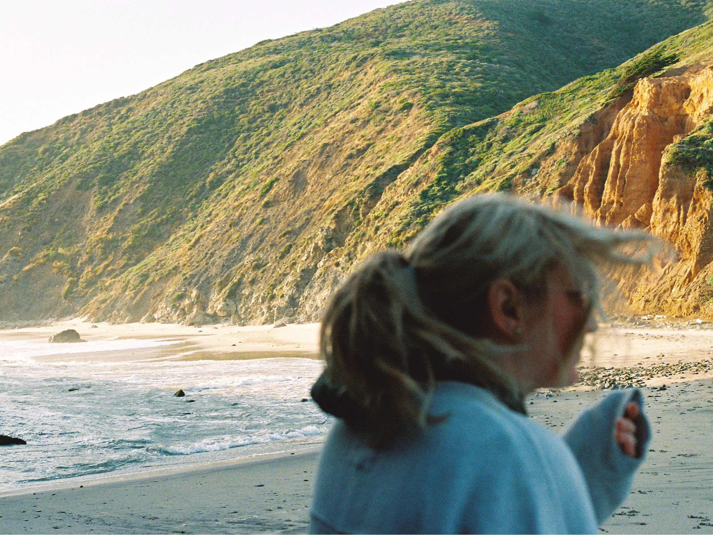 A woman with blonde hair, wearing glasses and a white sweater, standing on a beach with cliffs in the background, looking at her phone.