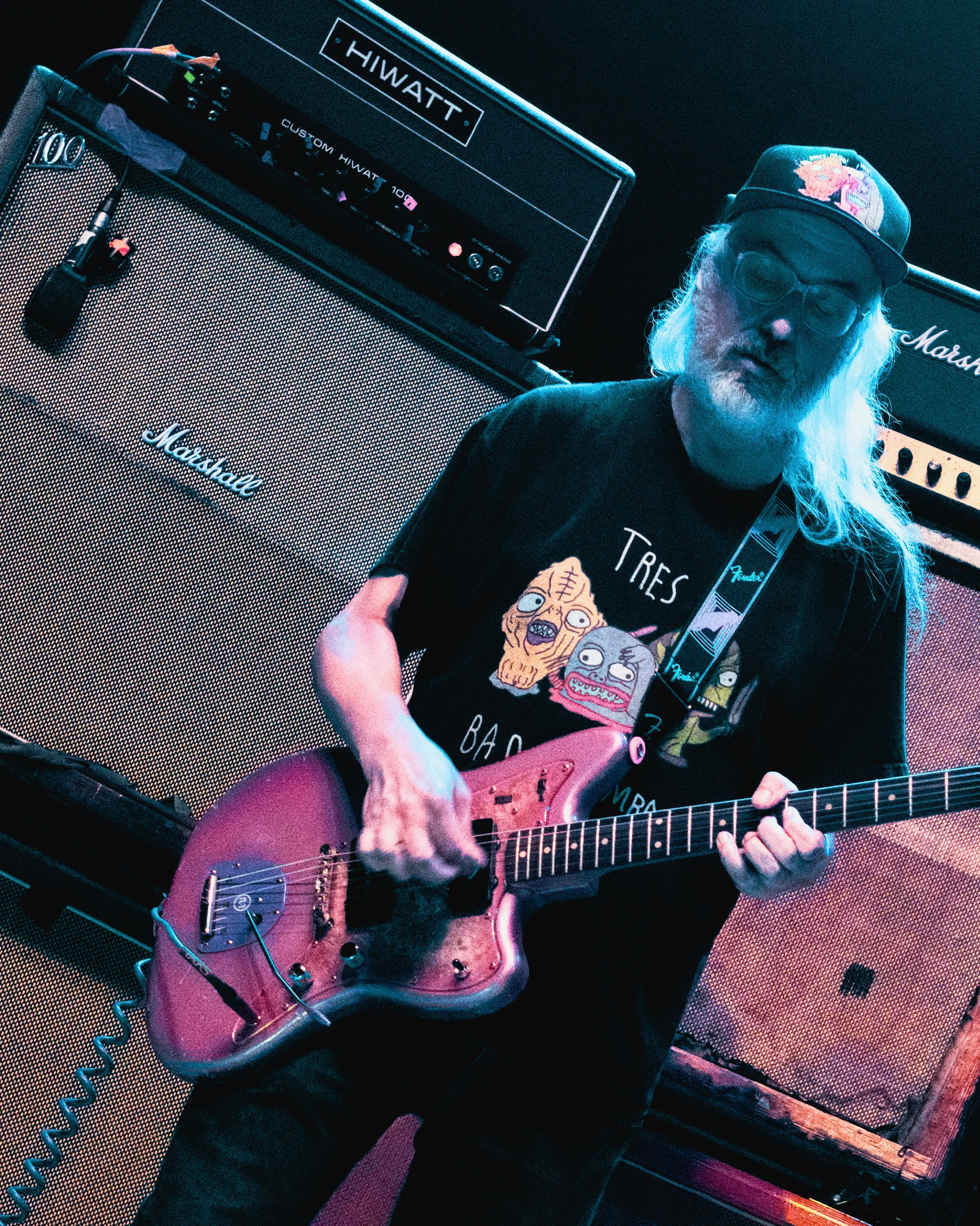 An elderly man with long white hair and beard playing a pink electric guitar in a music studio. Behind him are large Marshall amplifiers and a custom Hiwatt amplifier.