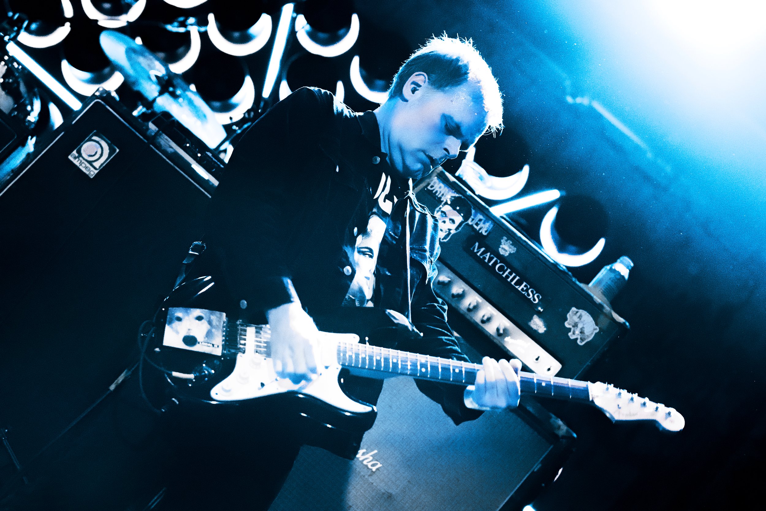 A man playing an electric guitar on stage, illuminated by blue stage lighting, with musical equipment and neon decorations in the background.