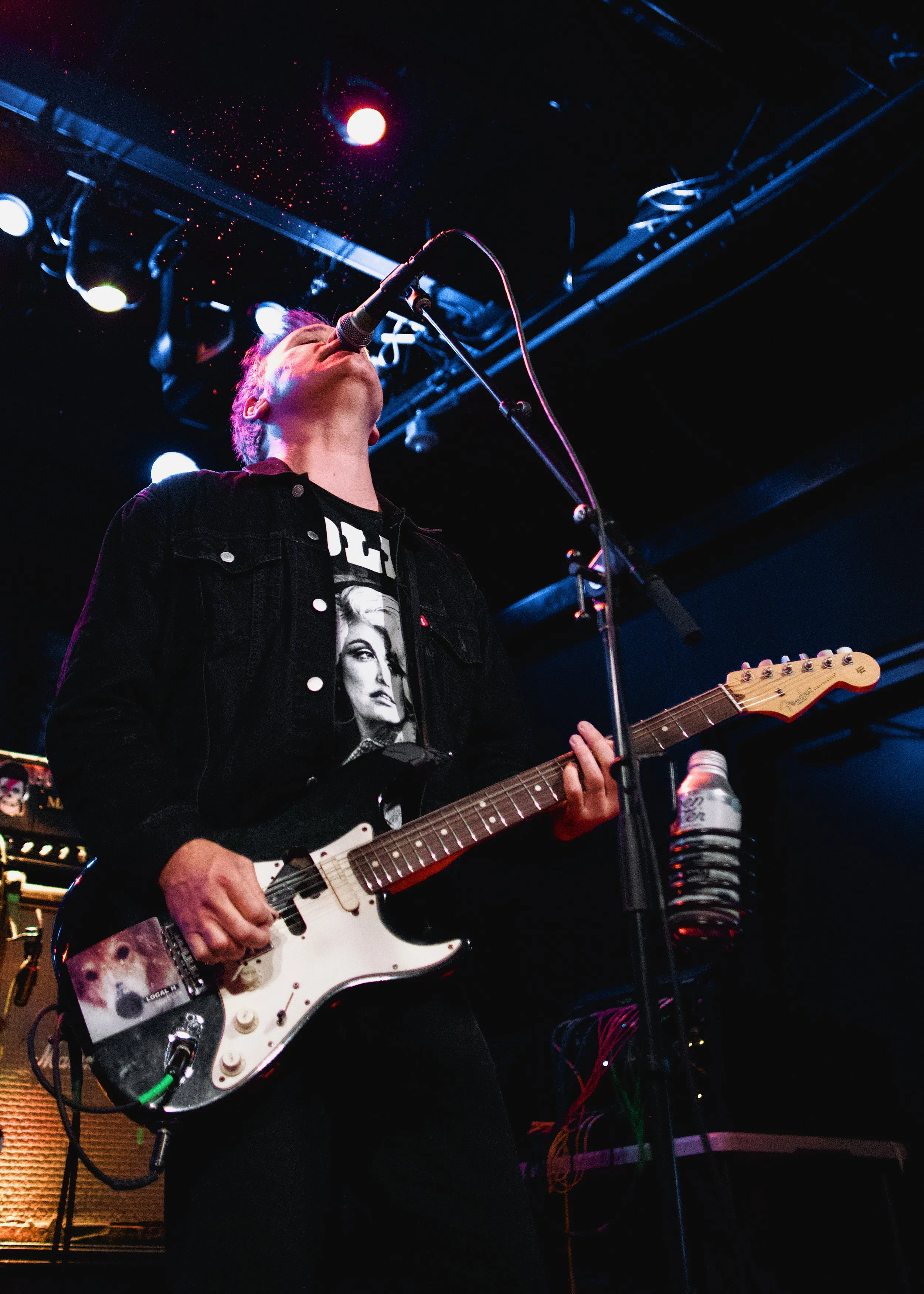A musician performing on stage with a black electric guitar, singing into a microphone, wearing a black jacket with a Marilyn Monroe graphic, and surrounded by stage lights and equipment.