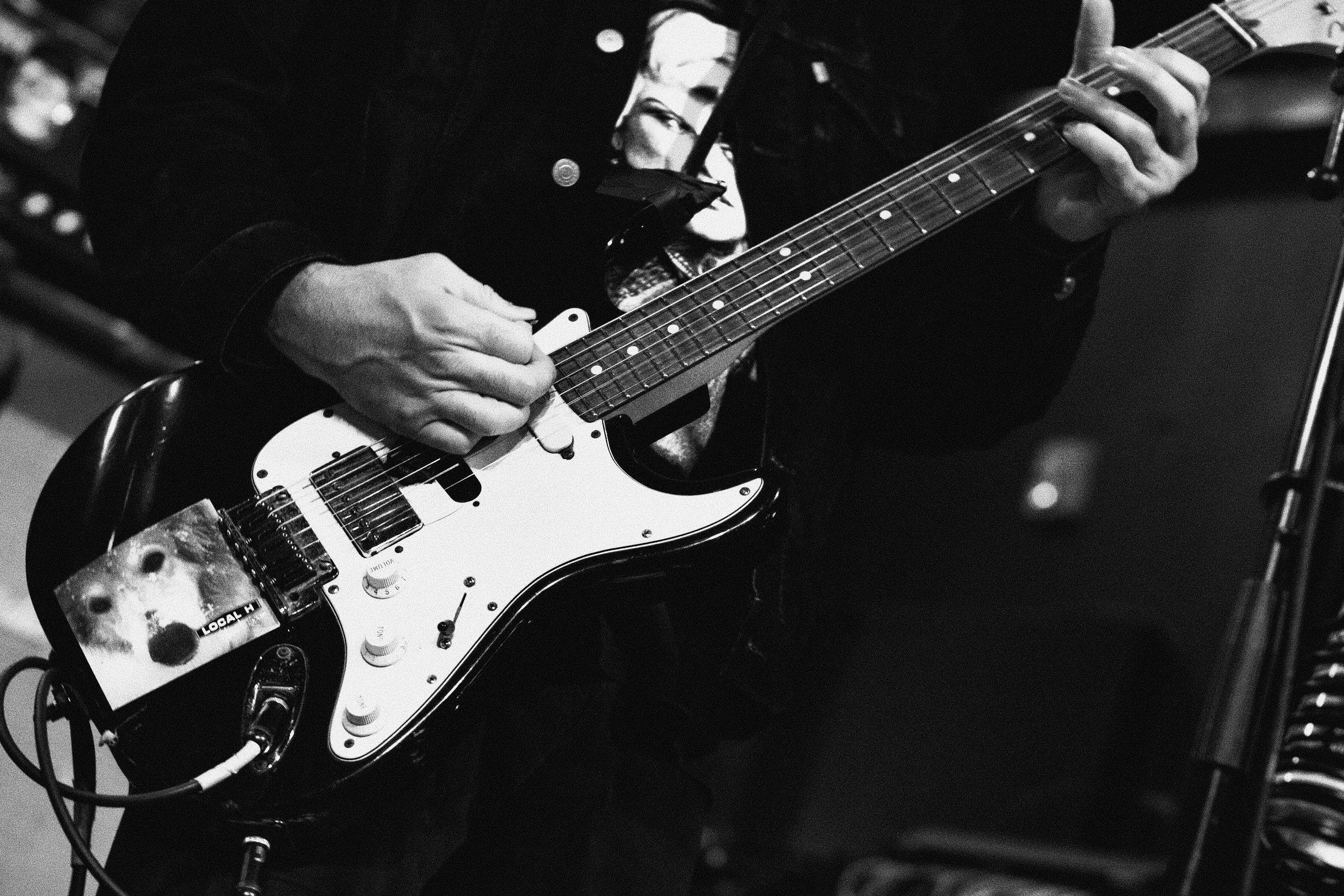 Close-up of a person playing an electric guitar, focusing on their hands and the guitar, in black and white.