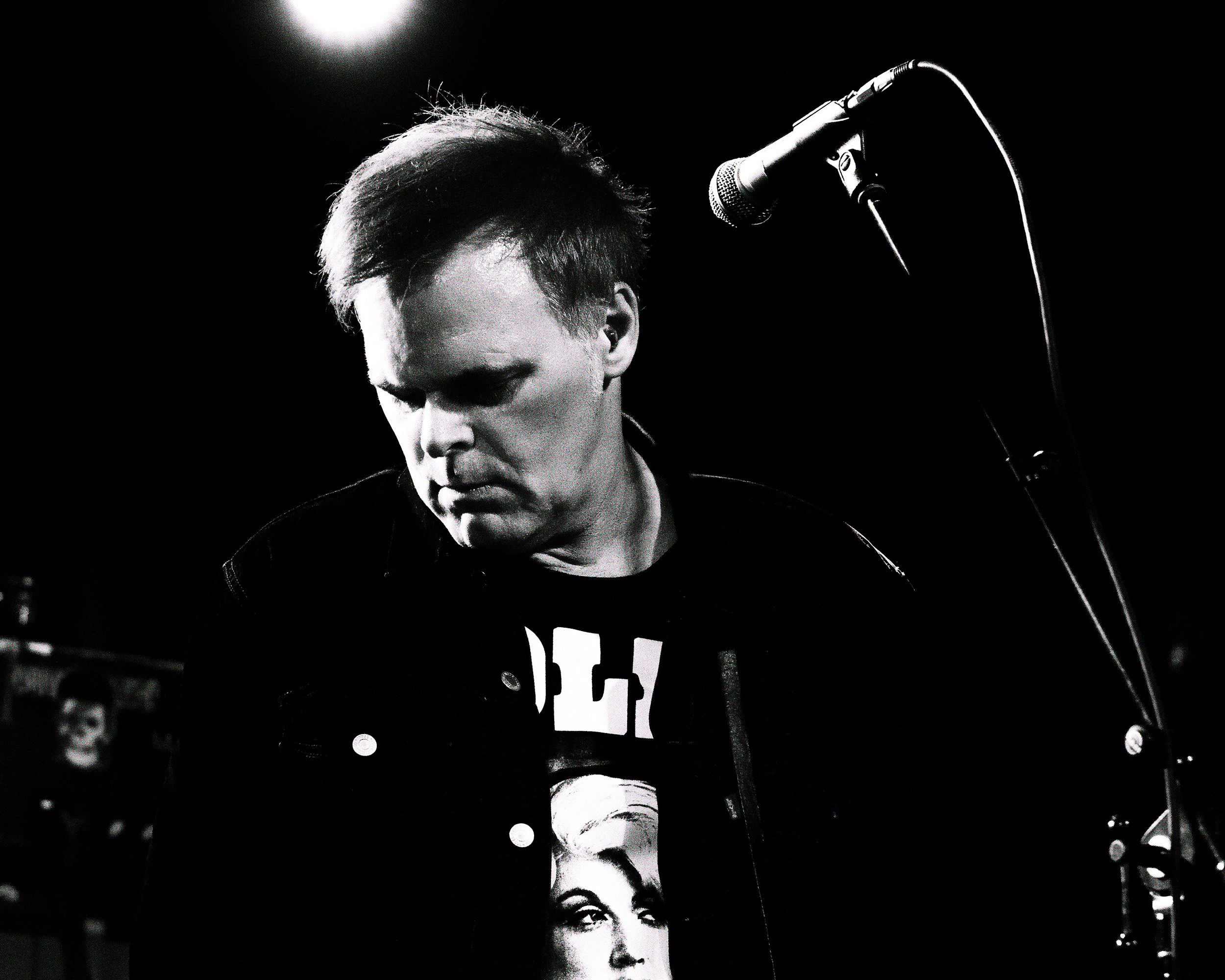 Black and white photo of a young man with short hair, looking down, standing near a microphone on a stand, in a dimly lit setting.