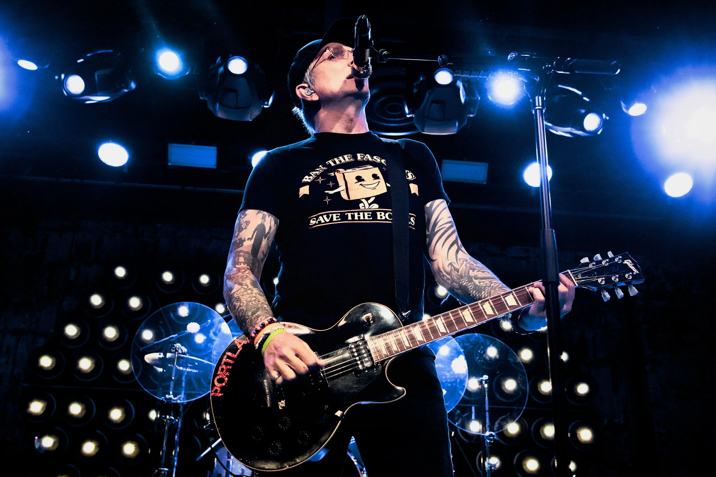 A musician performing on stage with a guitar, wearing a black t-shirt, glasses, and a black cap, surrounded by stage lights.