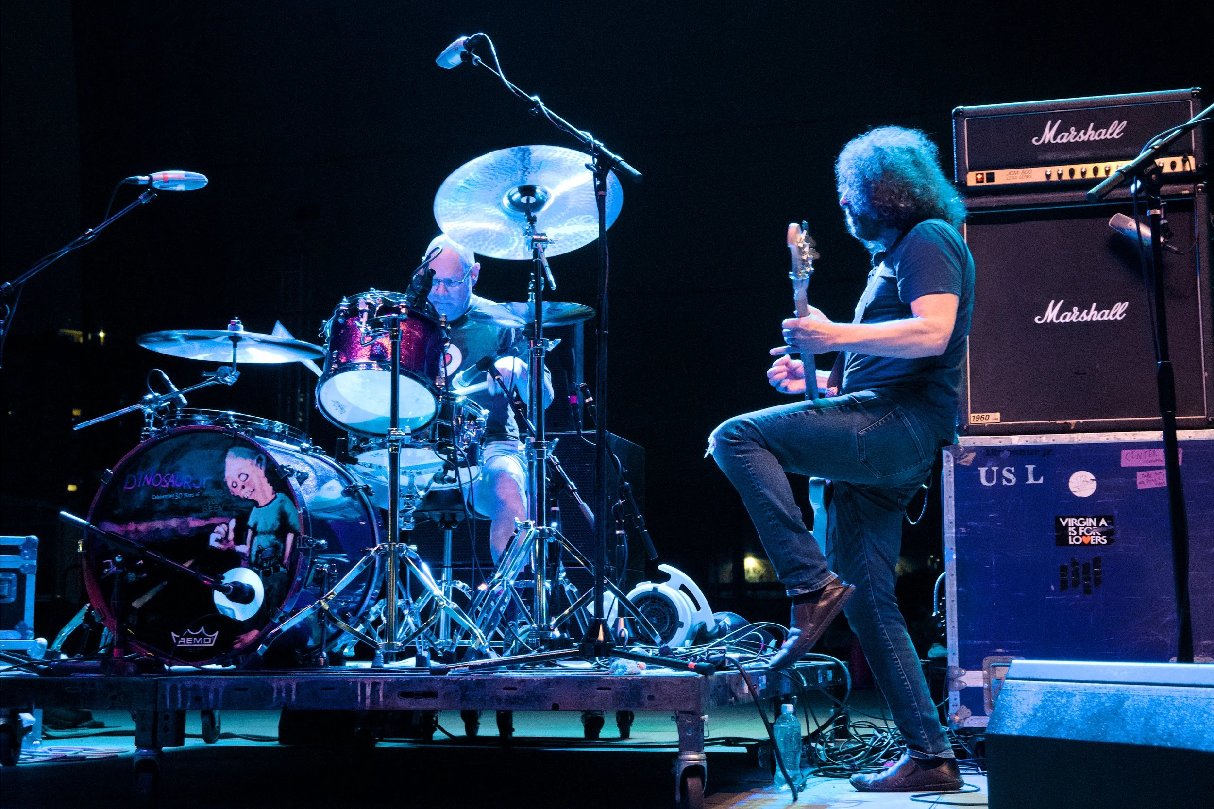 Two musicians perform on stage: one playing drums with a guitar pick, with a drumhead featuring a cartoon character, and the other playing an electric guitar near Marshall amplifiers. The stage is set with microphones and various equipment.