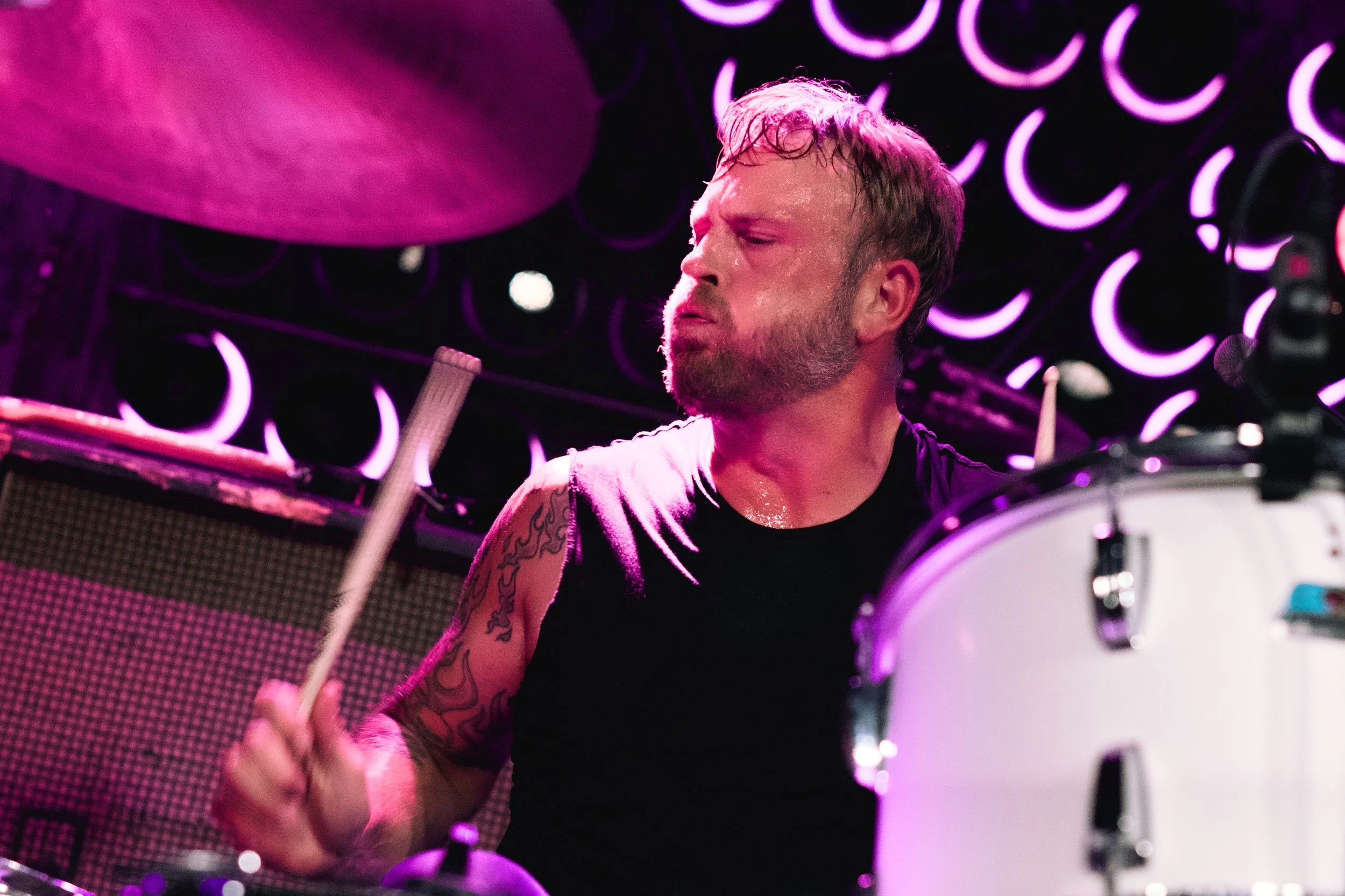 A man with a beard and tattooed arm playing drums on stage, illuminated by pink and purple stage lights, with neon crescent moon designs in the background.