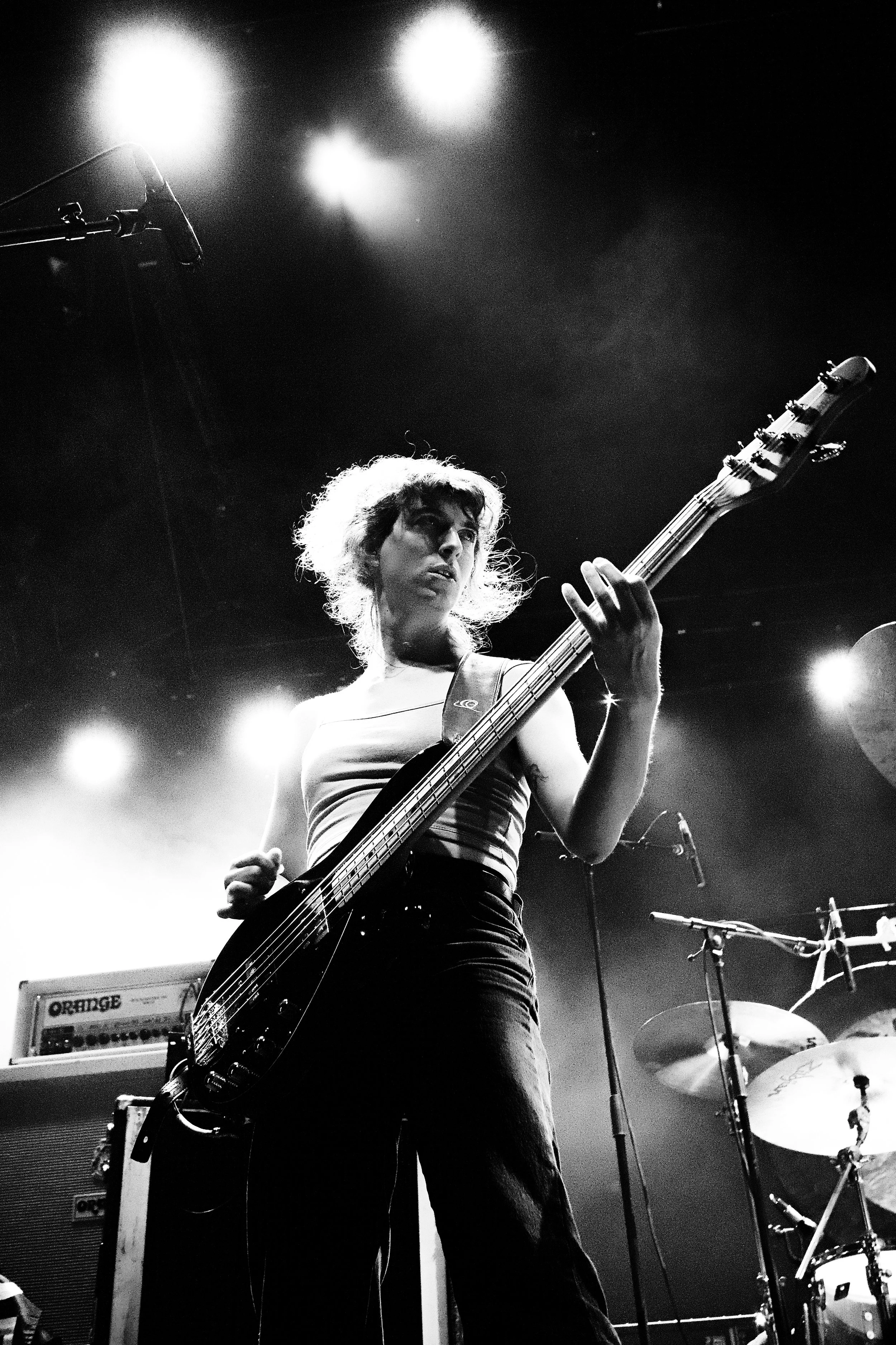 A female musician playing an electric bass guitar during a live concert, with stage lights illuminating her and a drum set visible in the background.