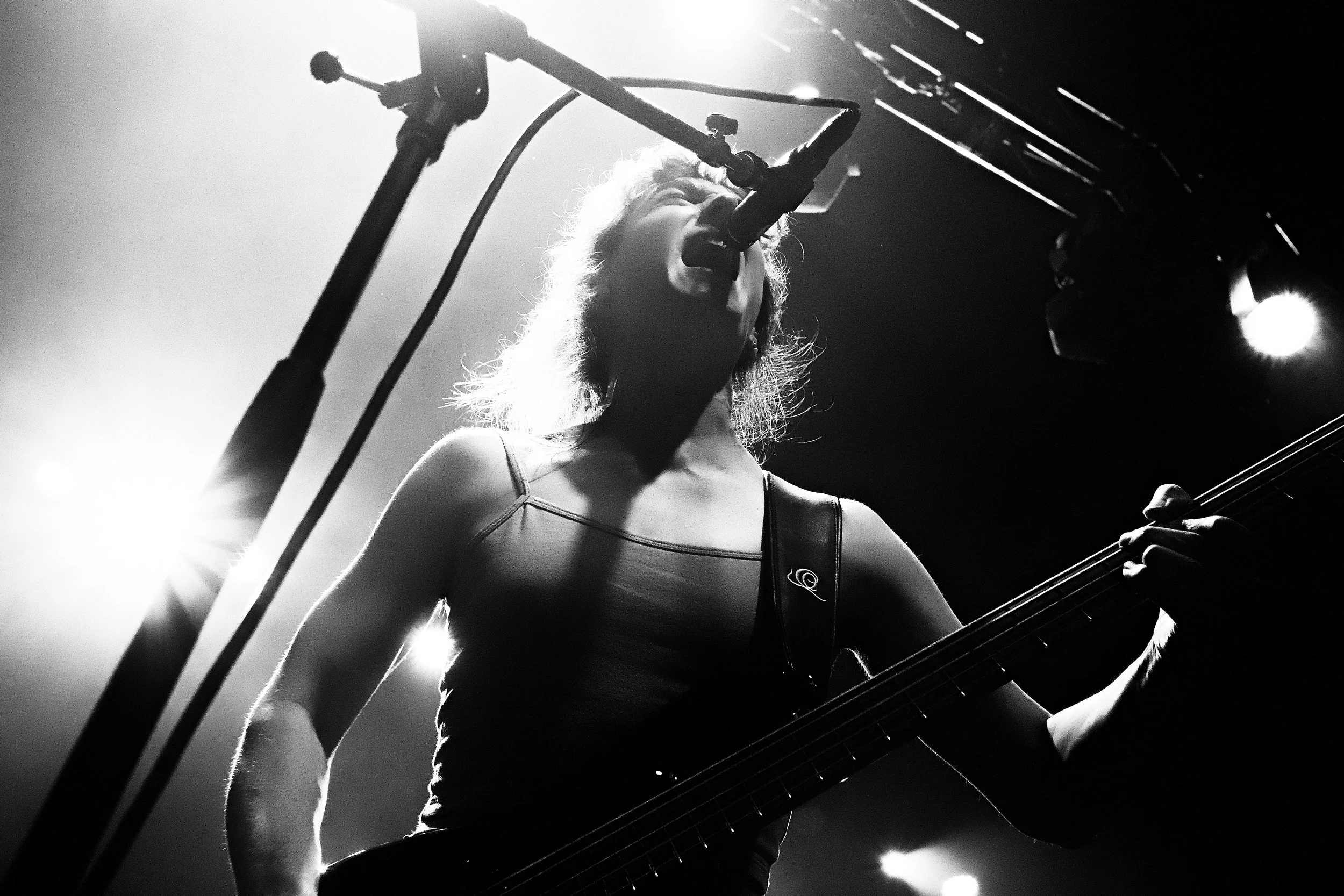 A female musician performs on stage, singing into a microphone and playing a guitar, with stage lights illuminating her from behind.