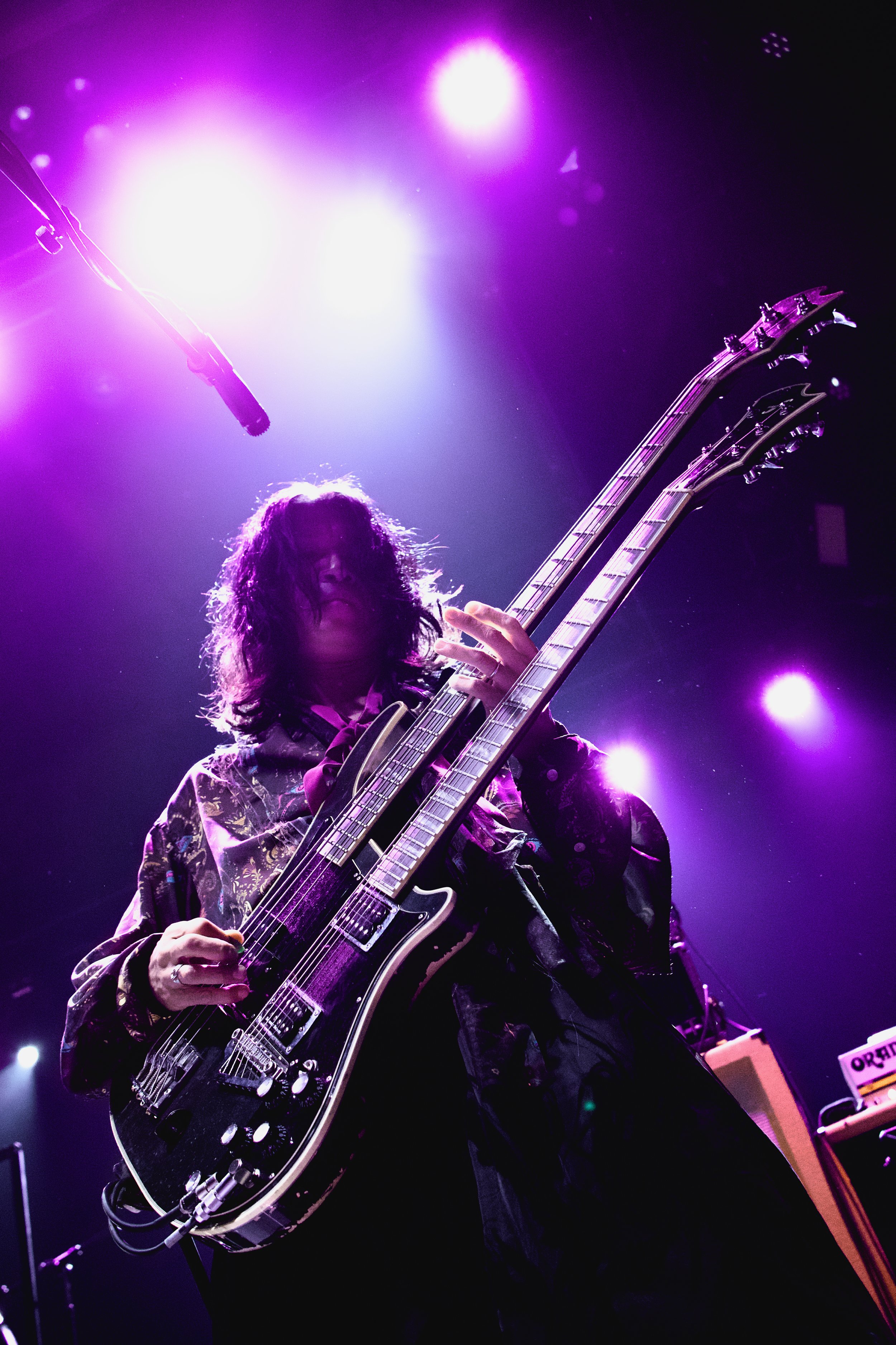 A musician with long curly hair playing a double-neck electric guitar on stage, illuminated by colorful purple and pink stage lights.