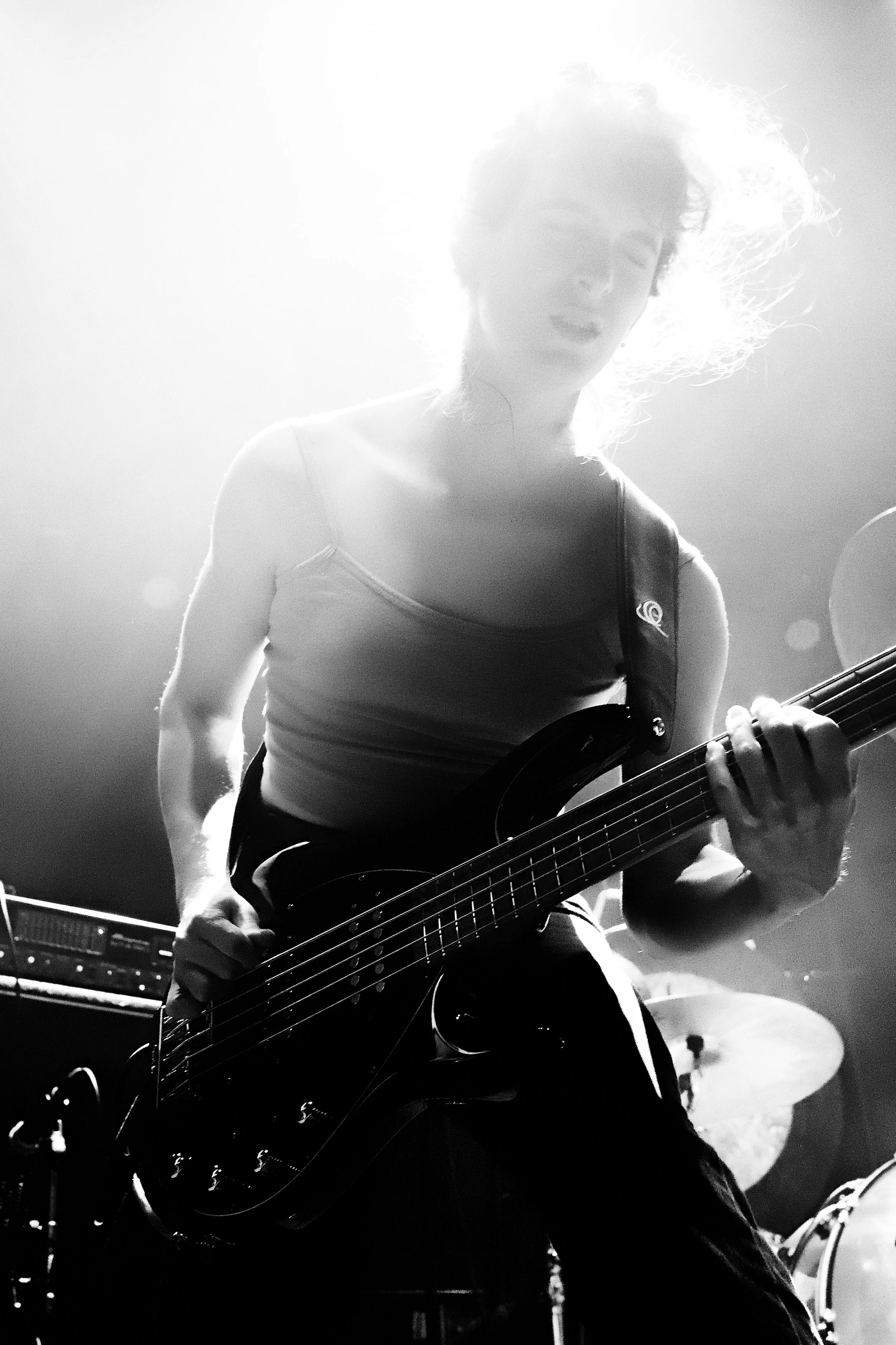 A female musician playing an electric bass guitar during a live performance, with bright stage lighting behind her.