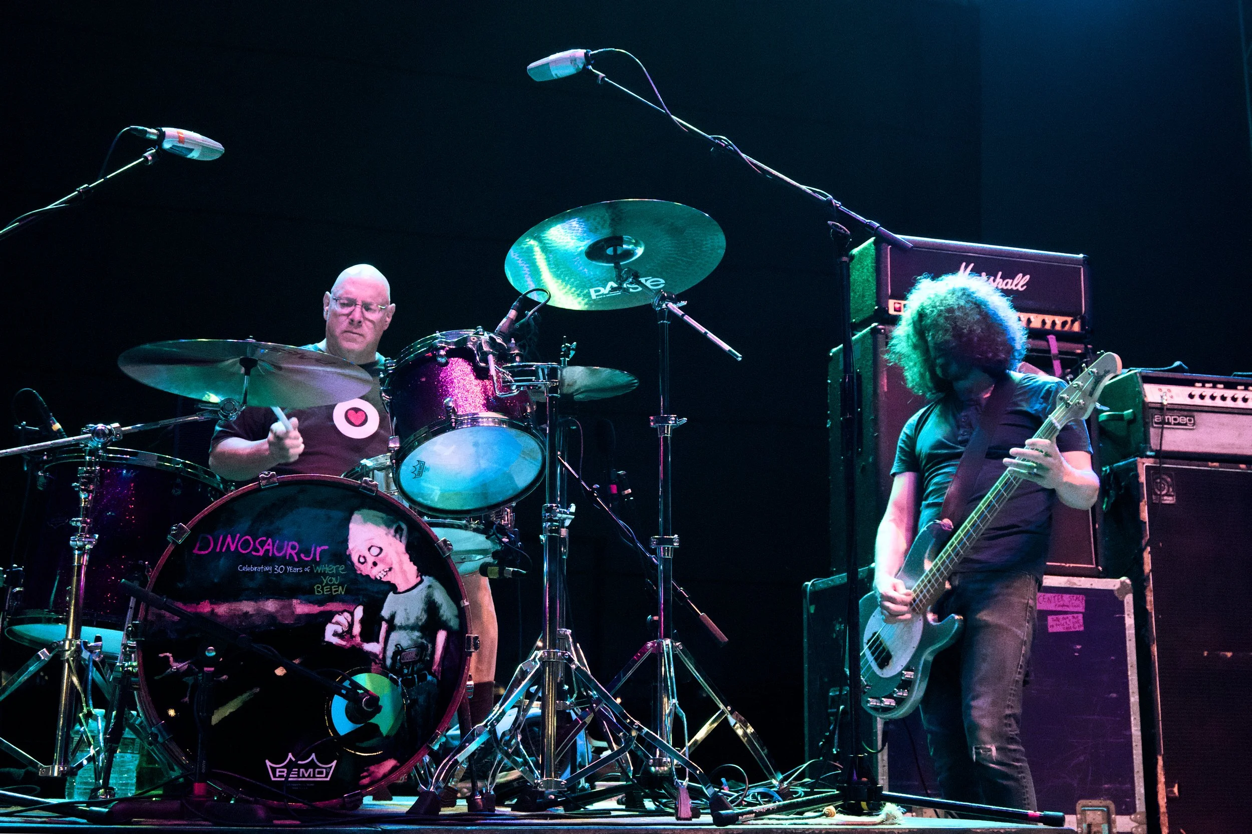 Two musicians performing on stage: one playing a drum set with a colorful bass drum featuring a skeleton and the words 'DINOSAUR JR,' the other playing an electric guitar next to large amplifiers, all under stage lighting.