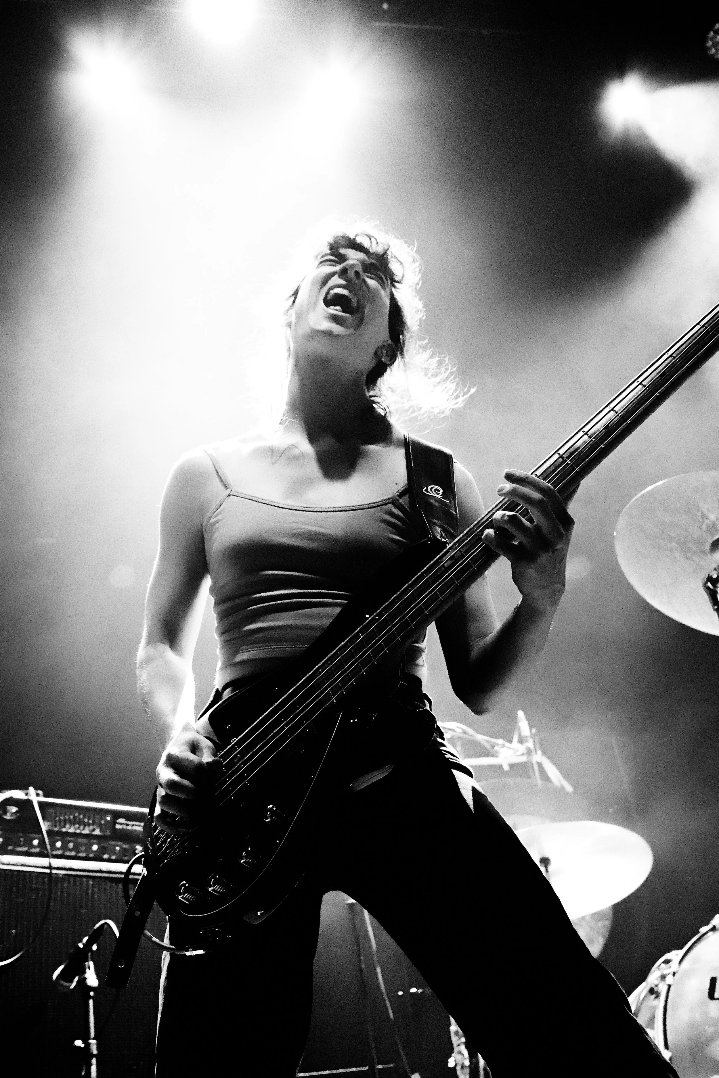 A black-and-white photo of a female musician playing a bass guitar passionately on stage, with her mouth open and eyes closed, illuminated by stage lights.