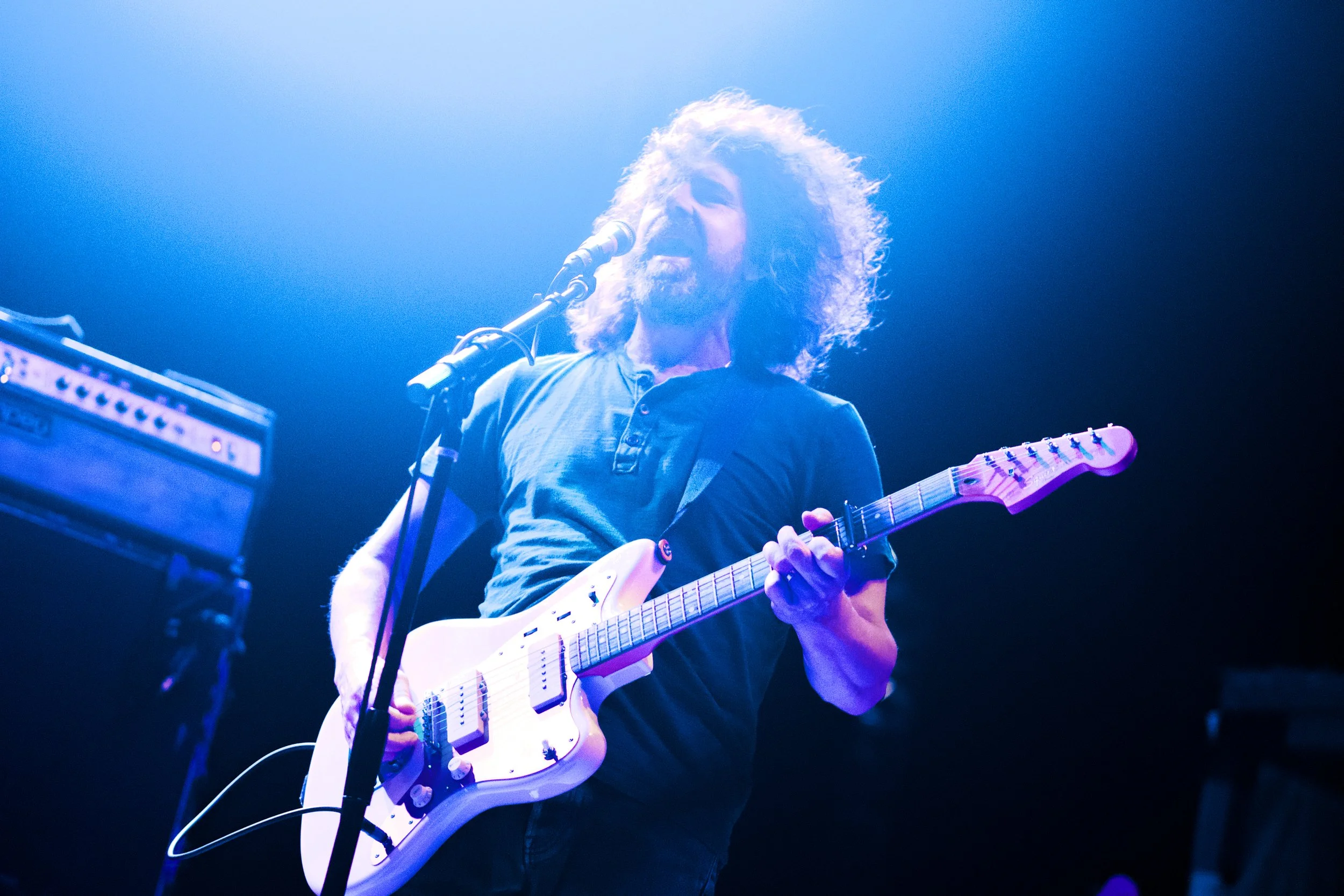 A male musician with curly hair playing an electric guitar on stage under blue lighting.