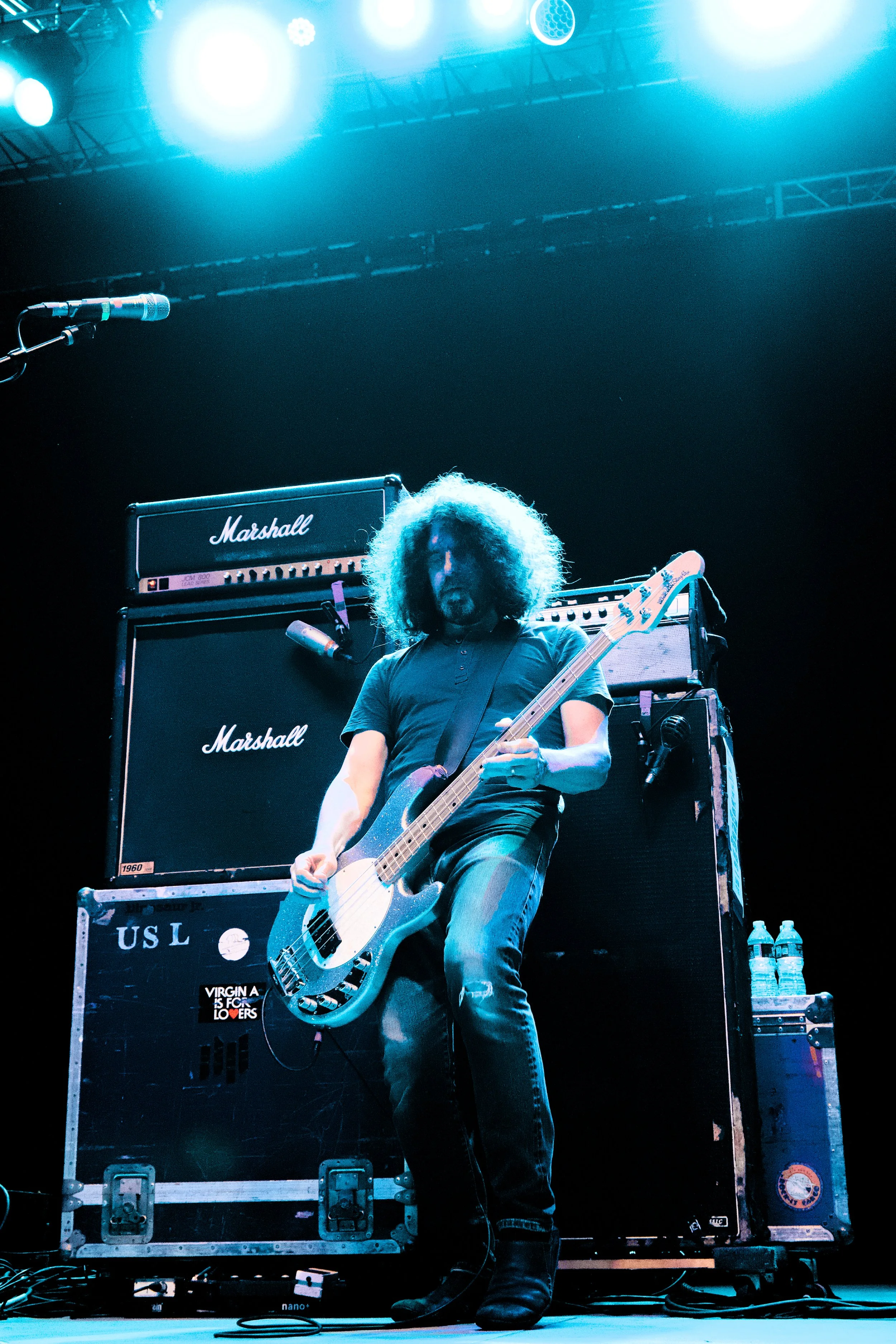 A musician with curly hair playing an electric bass guitar on stage, standing in front of large Marshall amplifiers with a microphone nearby, illuminated by bright stage lights.
