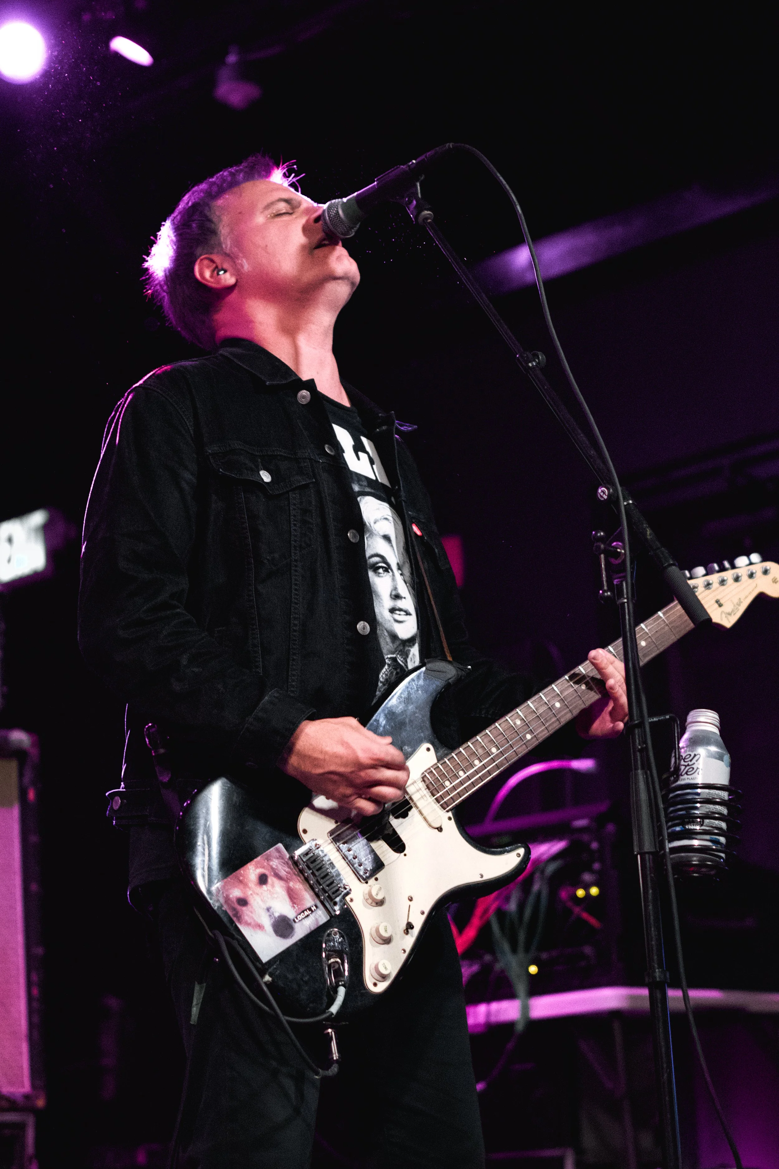 A male musician singing into a microphone and playing an electric guitar on stage. The stage is lit with purple and black lighting, and a water bottle is on the microphone stand.