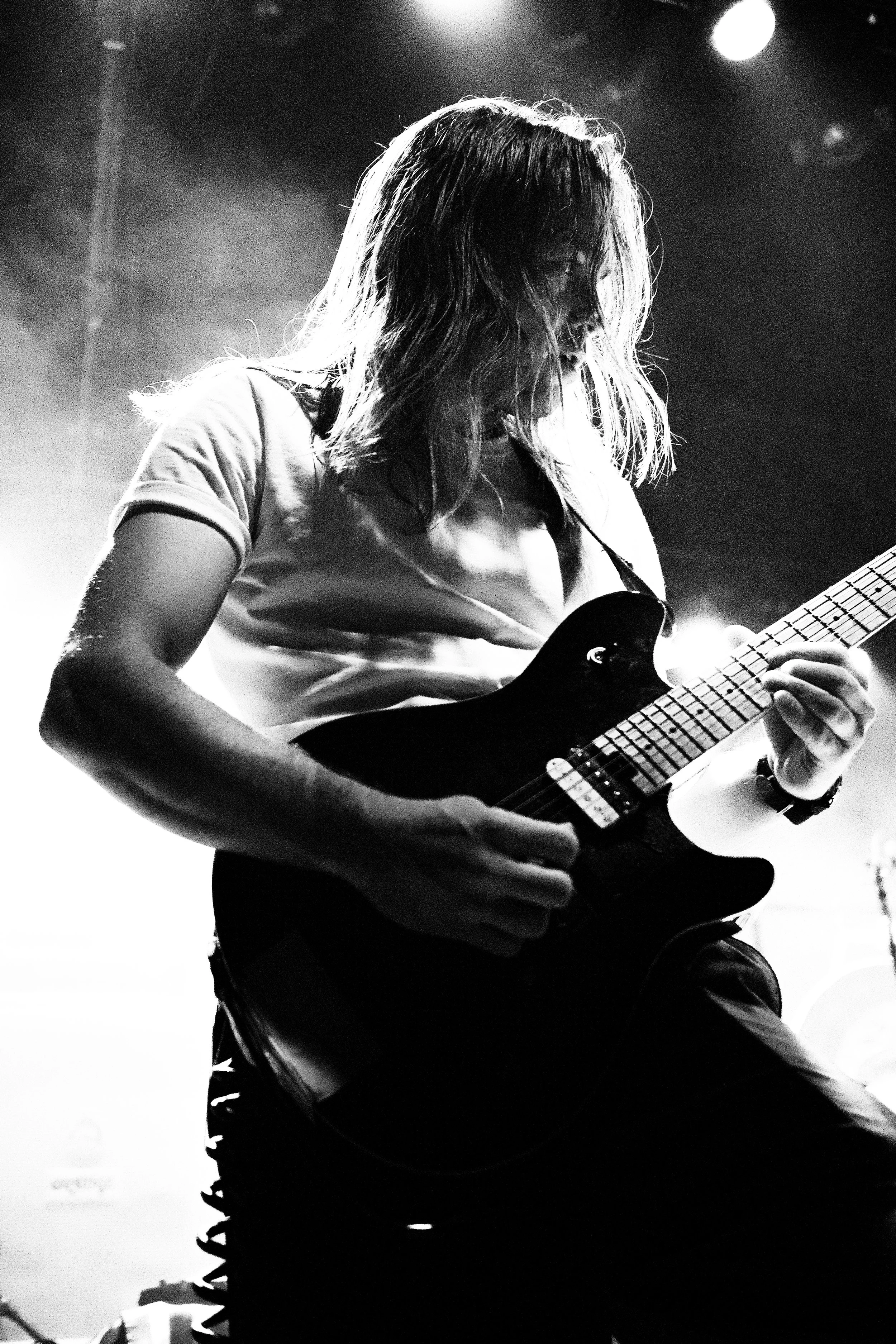 Black and white photo of a person with long hair playing an electric guitar on stage, with dramatic lighting.
