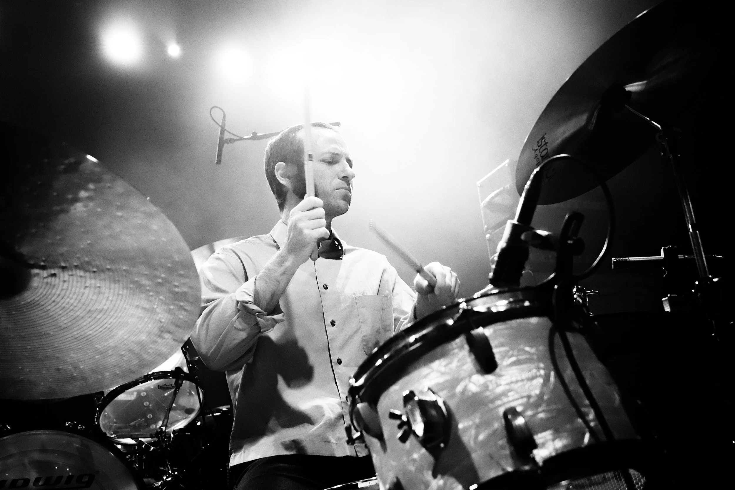 A man playing drums during a live music performance, with stage lights shining overhead.
