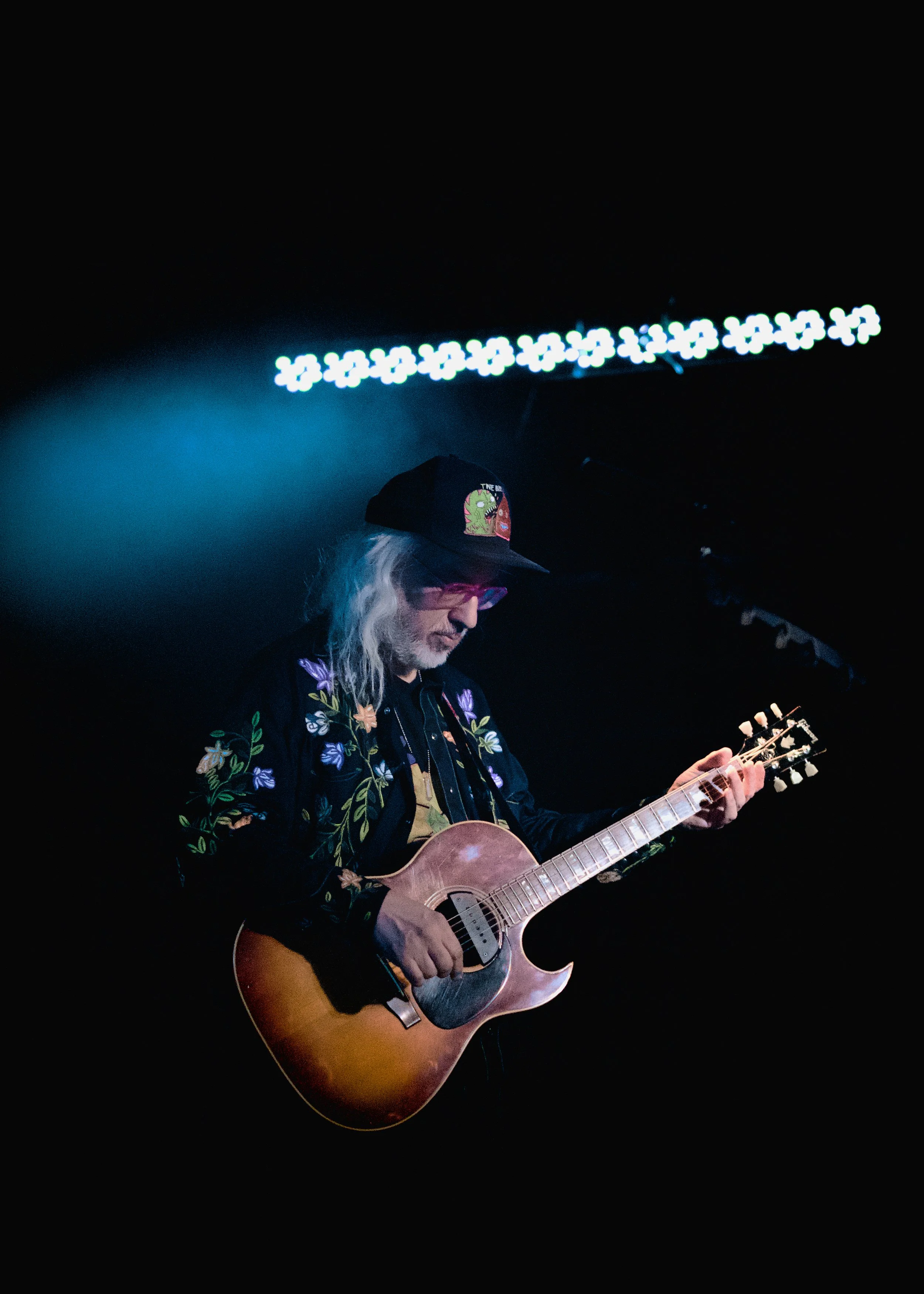 A musician with long gray hair and glasses playing an acoustic guitar on stage, wearing a black cap with colorful patches, a black embroidered jacket, illuminated by stage lights.