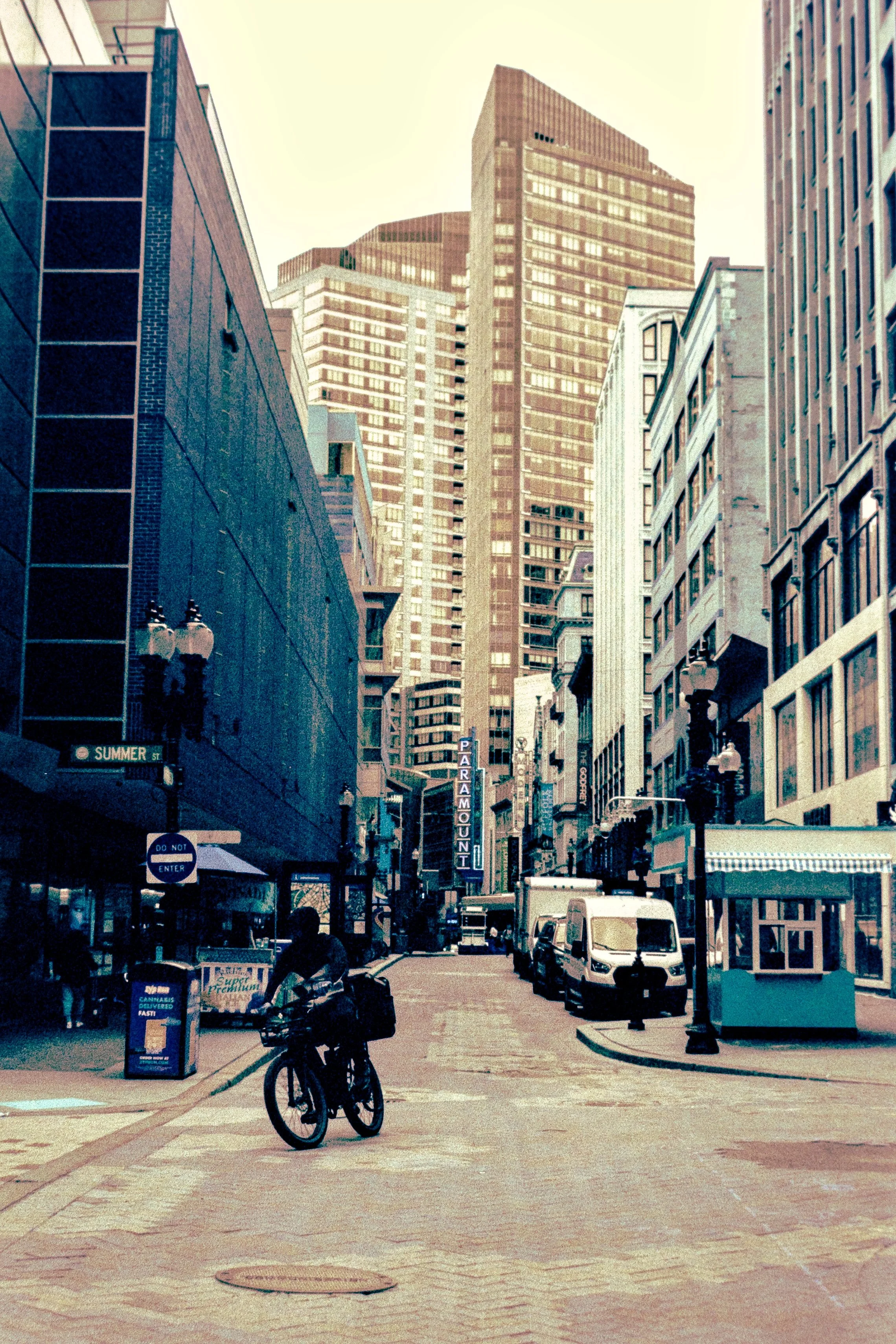 A city street with tall buildings, a cyclist, parked vehicles, and a vintage theater sign that reads 'Paramount'.