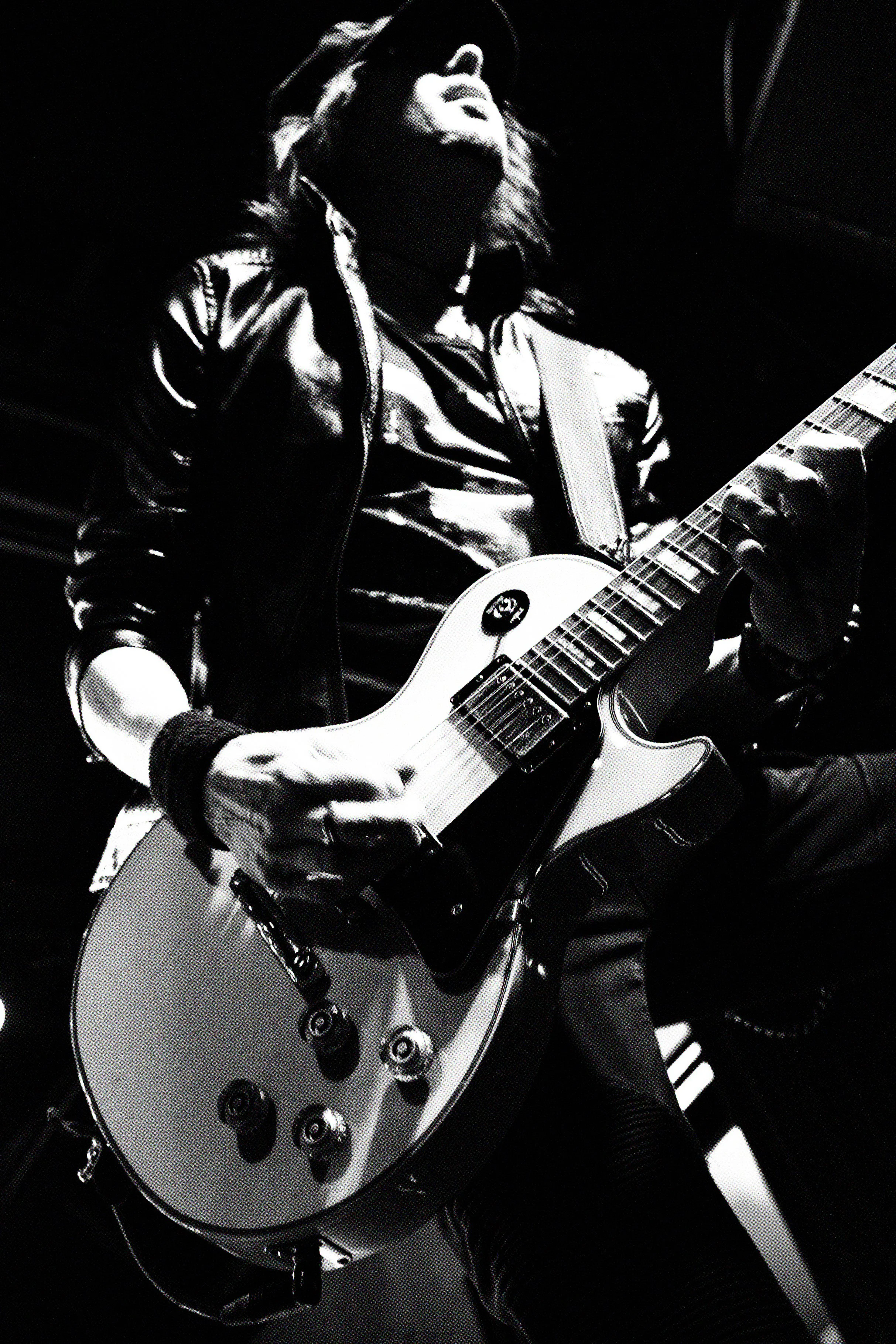 A person playing an electric guitar on stage, in black and white, wearing a leather jacket and a wristband, with long hair and a cap, looking upwards.