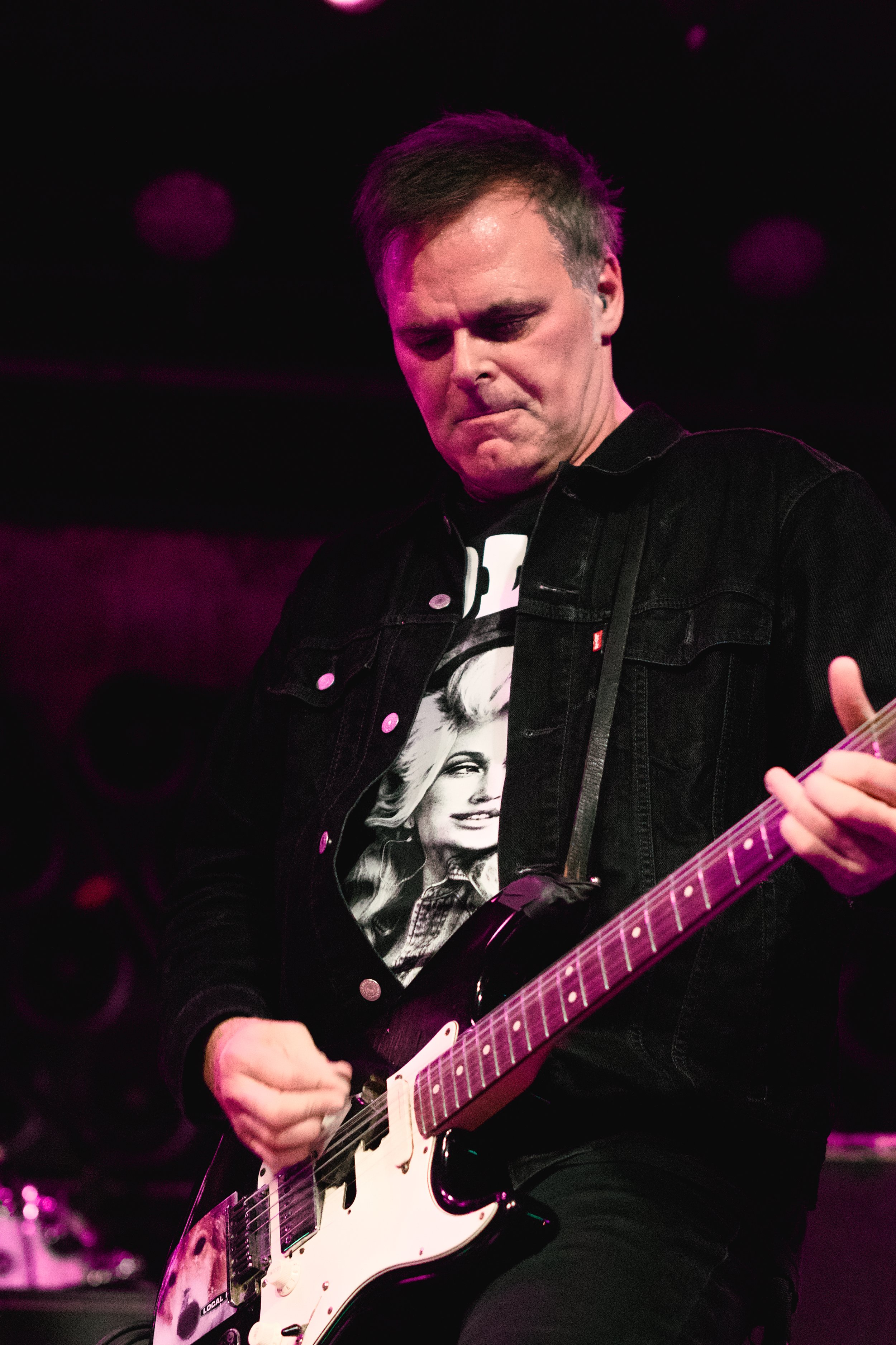 A man playing an electric guitar on stage, wearing a black jacket with a graphic t-shirt, with stage lights in pink and purple tones in the background.