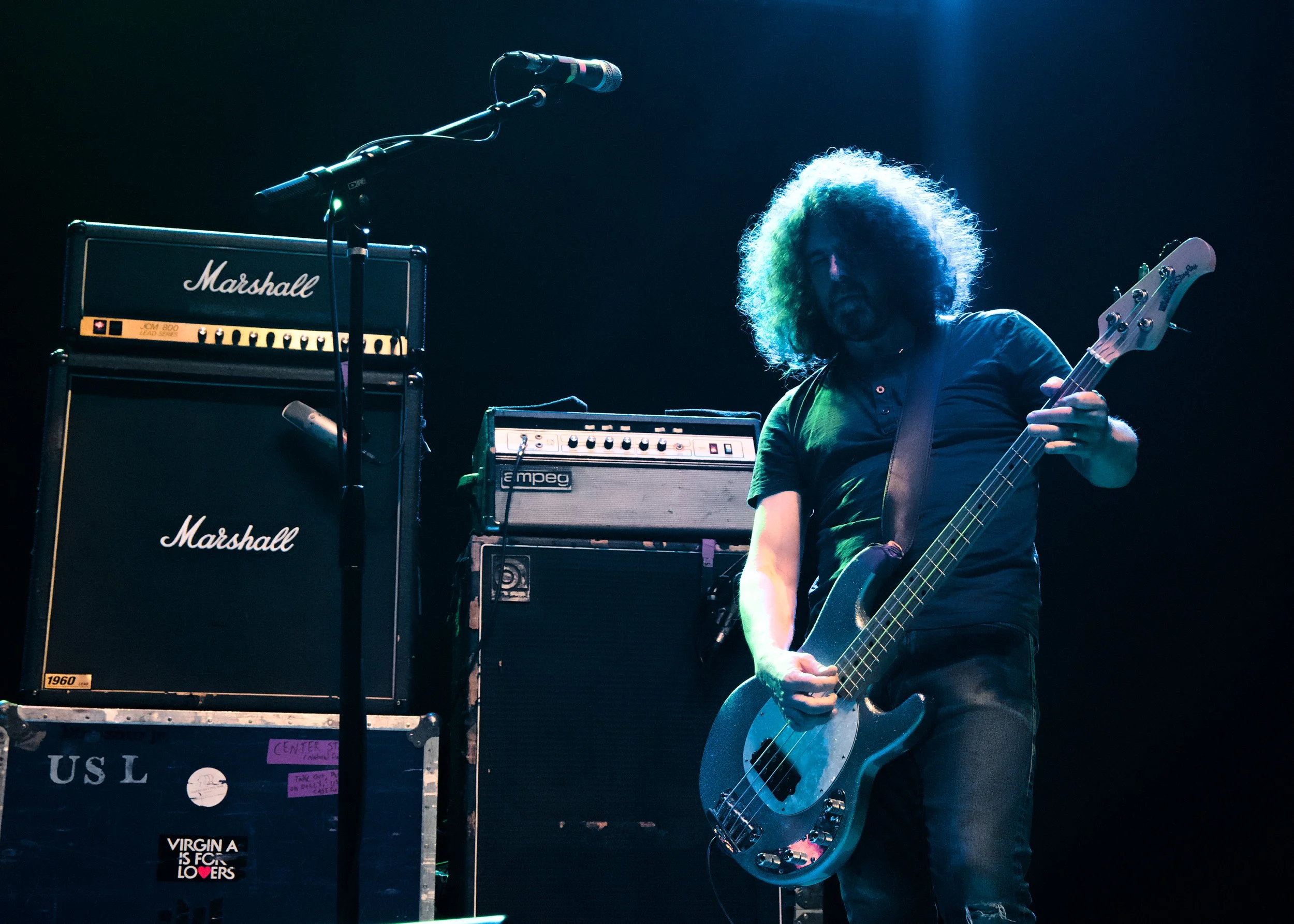 A man with curly hair playing an electric bass guitar on stage, surrounded by Marshall and Empeg amplifiers.